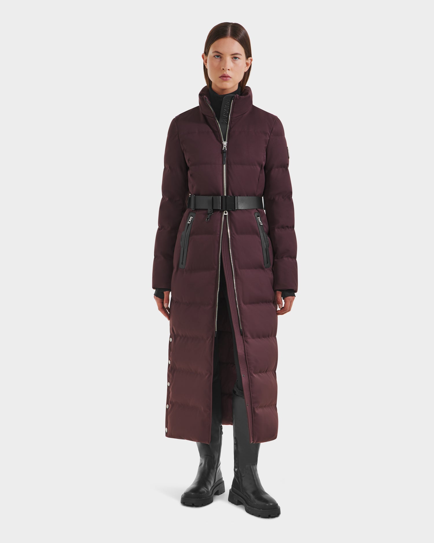 A woman faces forward wearing the Rudsak IZABELLA H - 8125644 Bordeaux, a long burgundy puffer coat with a belt, high collar, zippered pockets, water-resistant finish; styled with a black turtleneck and chunky boots against a white background.