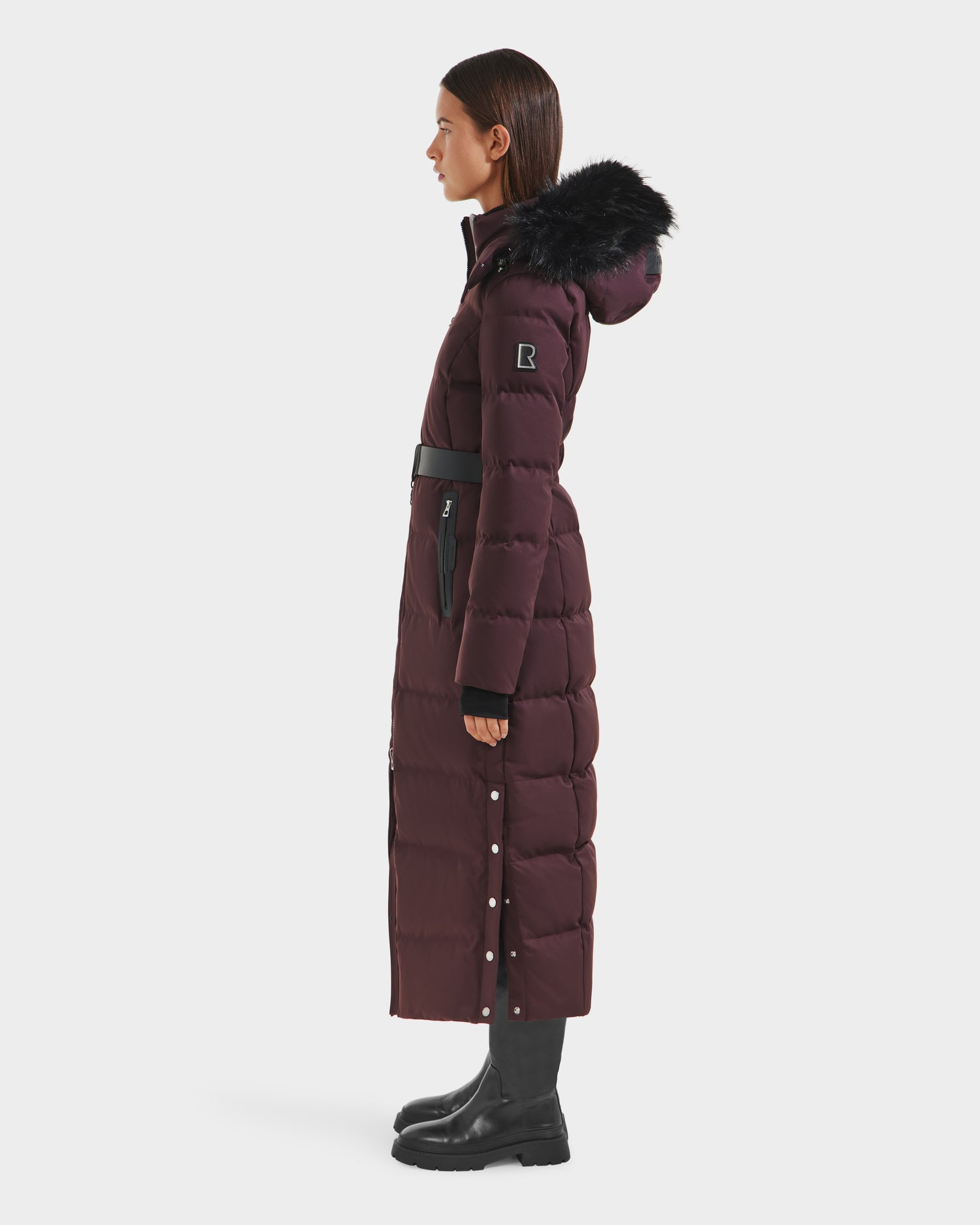 A woman stands in profile wearing the Rudsak IZABELLA H - 8125644 Bordeaux puffer down coat with a black fur-trimmed hood, belted waist, button side details, and water-resistant finish for style and practicality.