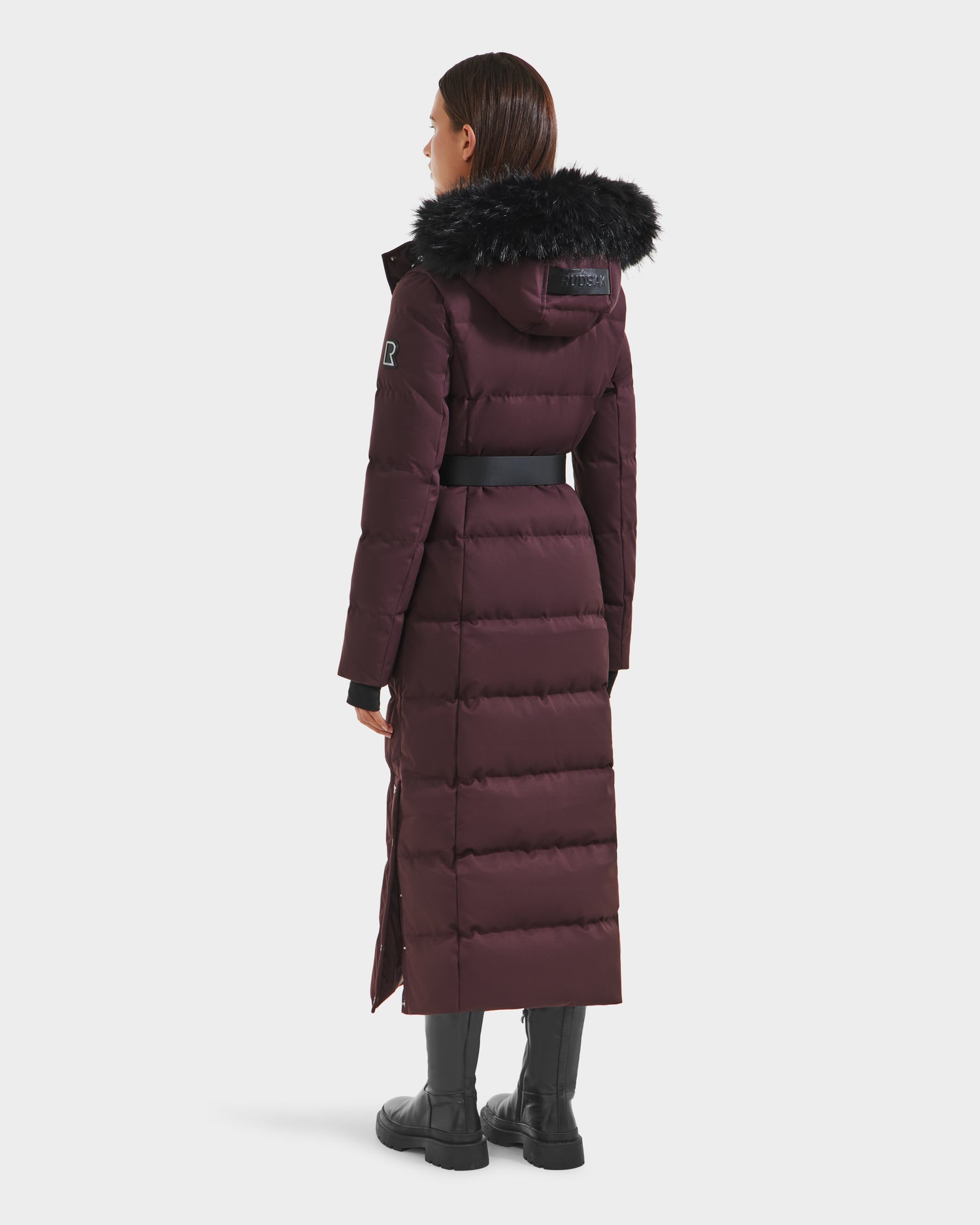 A woman faces away, wearing the Rudsak IZABELLA H - 8125644 Bordeaux women’s puffer down coat with black fur-trimmed hood, black belt, water-resistant fabric, horizontal quilting, and calf-length design.