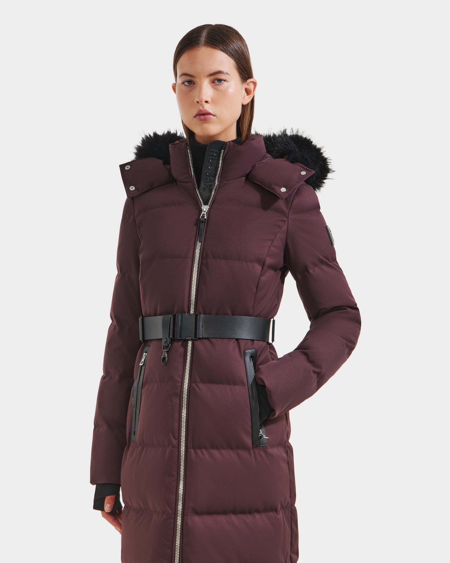 A woman models the Rudsak IZABELLA H - 8125644 Bordeaux, a long maroon puffer winter jacket with a black faux fur-trimmed hood and wide black belt, against a plain white background.