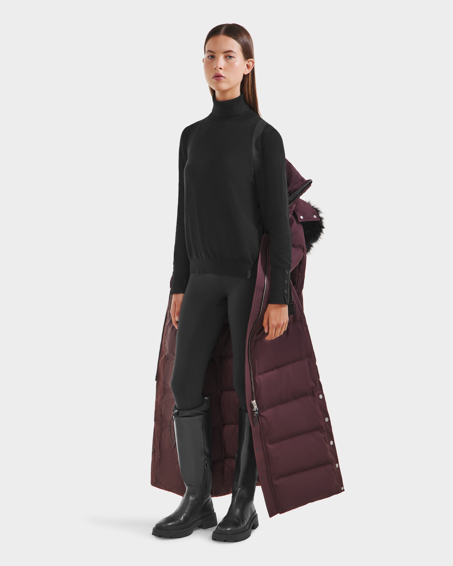 A woman in a black turtleneck, leggings, and boots has the Rudsak IZABELLA H - 8125644 Bordeaux maroon water-resistant puffer draped over her shoulder, looking straight ahead against a white background.