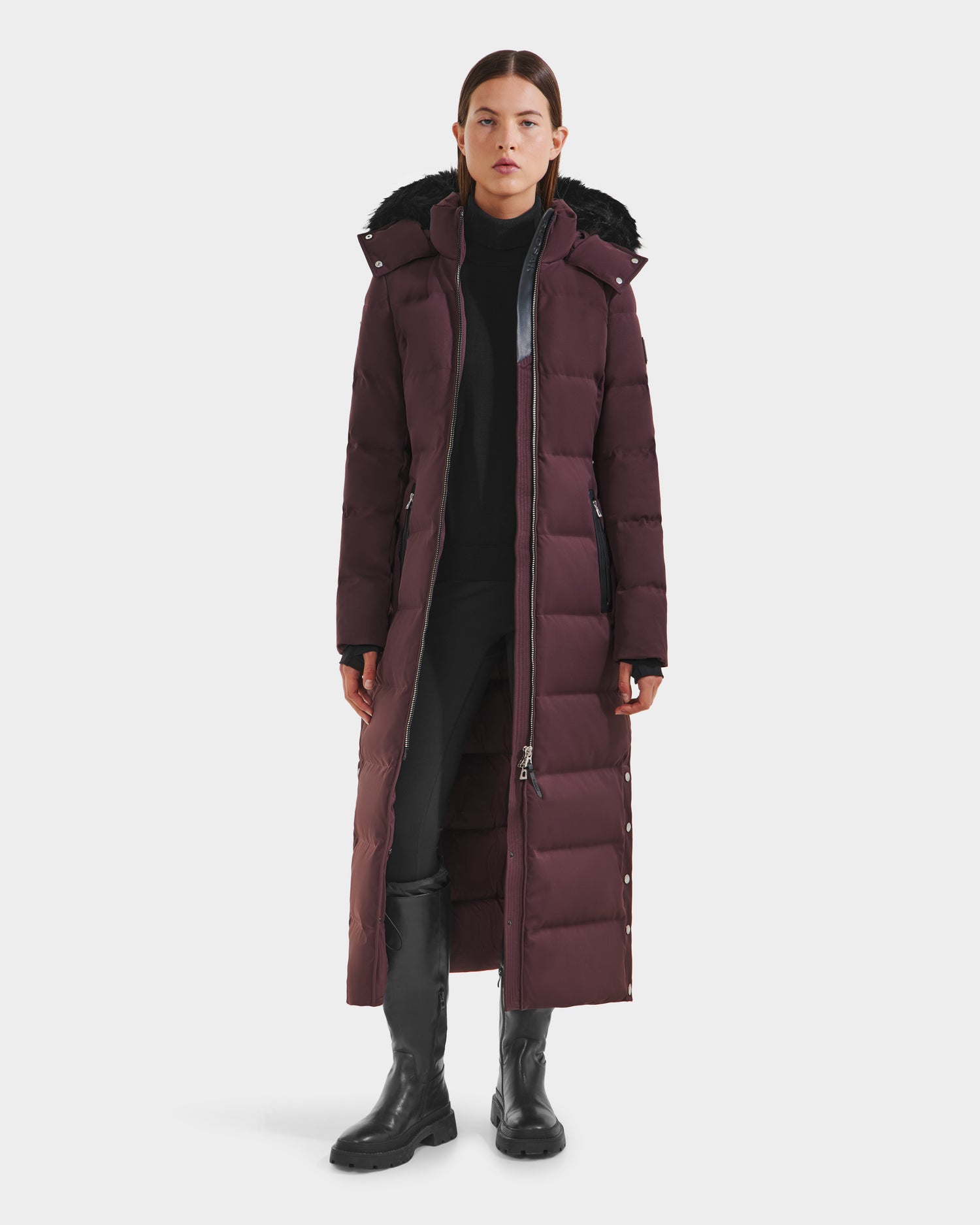 A woman faces forward wearing the Rudsak IZABELLA H - 8125644 Bordeaux, a long maroon water-resistant quilted coat with a hood, black pants, boots, and a black top visible beneath the buttoned and partially unzipped coat.