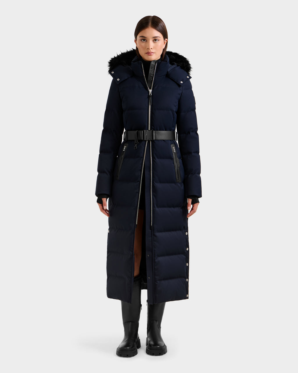 A woman stands facing forward wearing the Rudsak IZABELLA H - 8125644 Navy puffer cold-weather coat with RDS-certified down, black belt, fur-trimmed hood, and black boots against a plain light gray background.