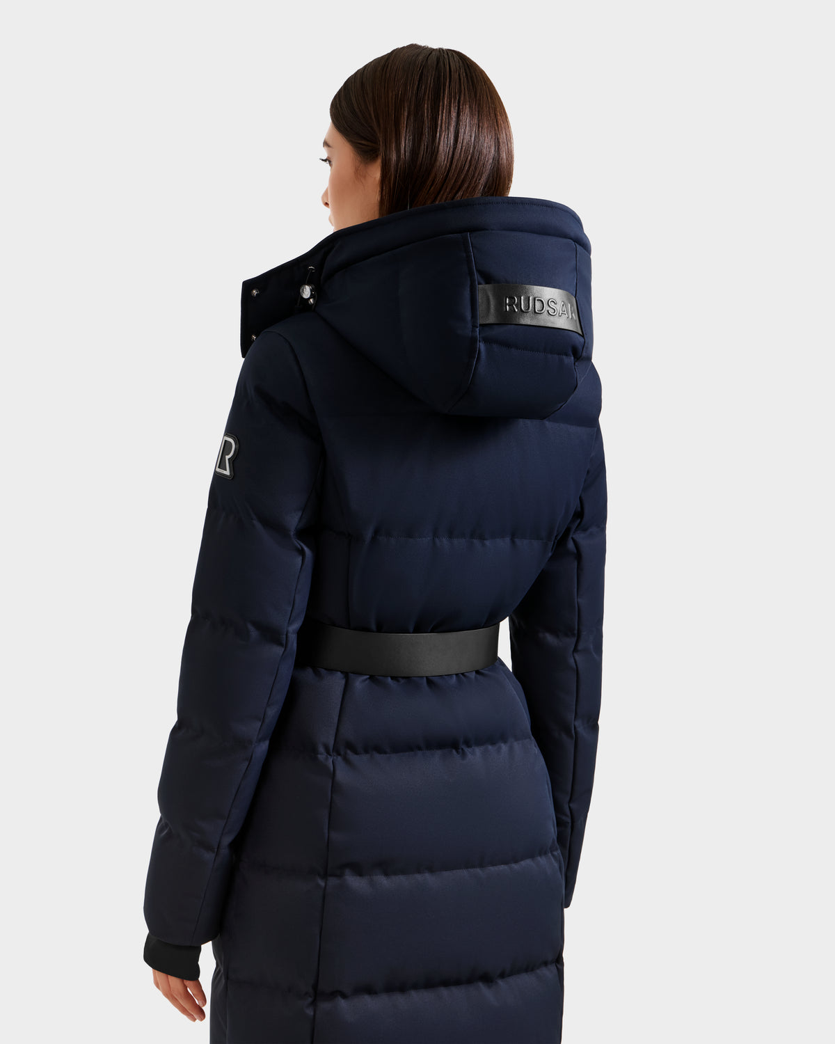 A woman, seen from the back, wears the Rudsak IZABELLA H - 8125644 Navy puffer coat with hood and black belt. The RDS-certified down coat features a Rudsak label on the hood and is designed for cold weather warmth.