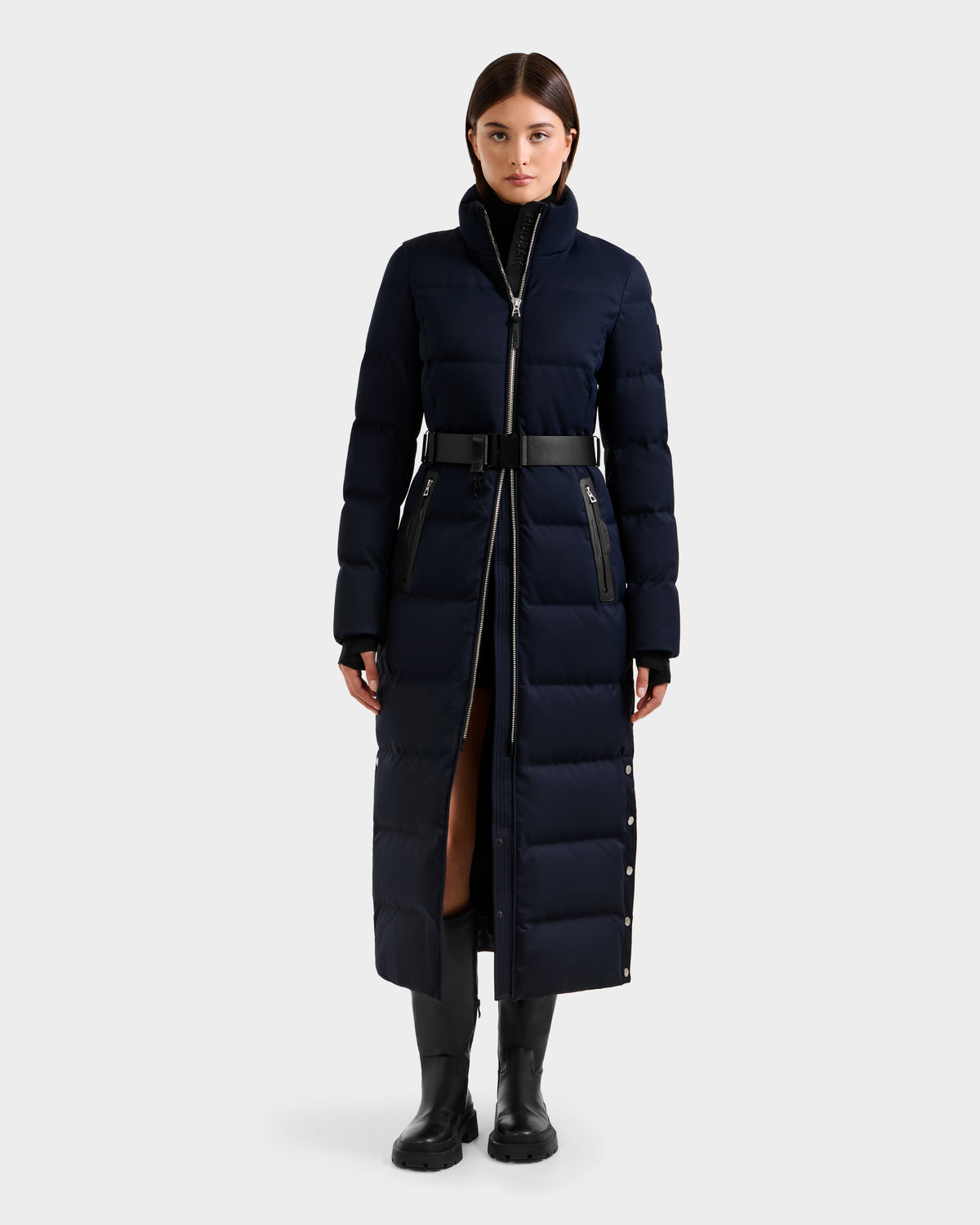 A woman faces forward against a light gray background, wearing the Rudsak IZABELLA H - 8125644 Navy puffer coat styled with a belt, paired with a black turtleneck, black pants, and black boots.
