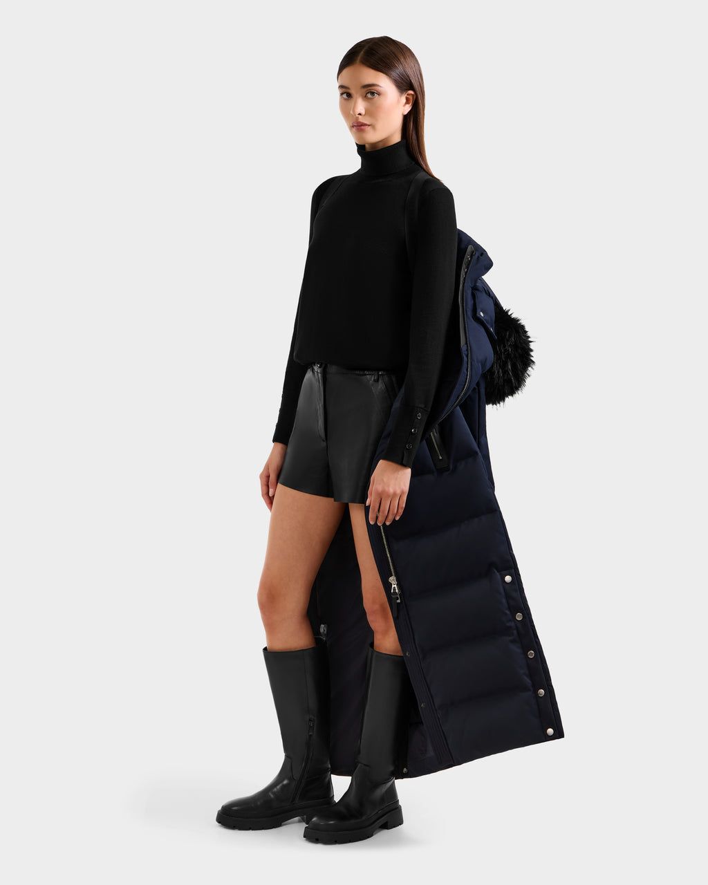 A woman in a black turtleneck, leather shorts, and knee-high boots holds the Rudsak IZABELLA H - 8125644 Navy women’s puffer coat with a fur-trimmed hood and RDS-certified down over her arm.