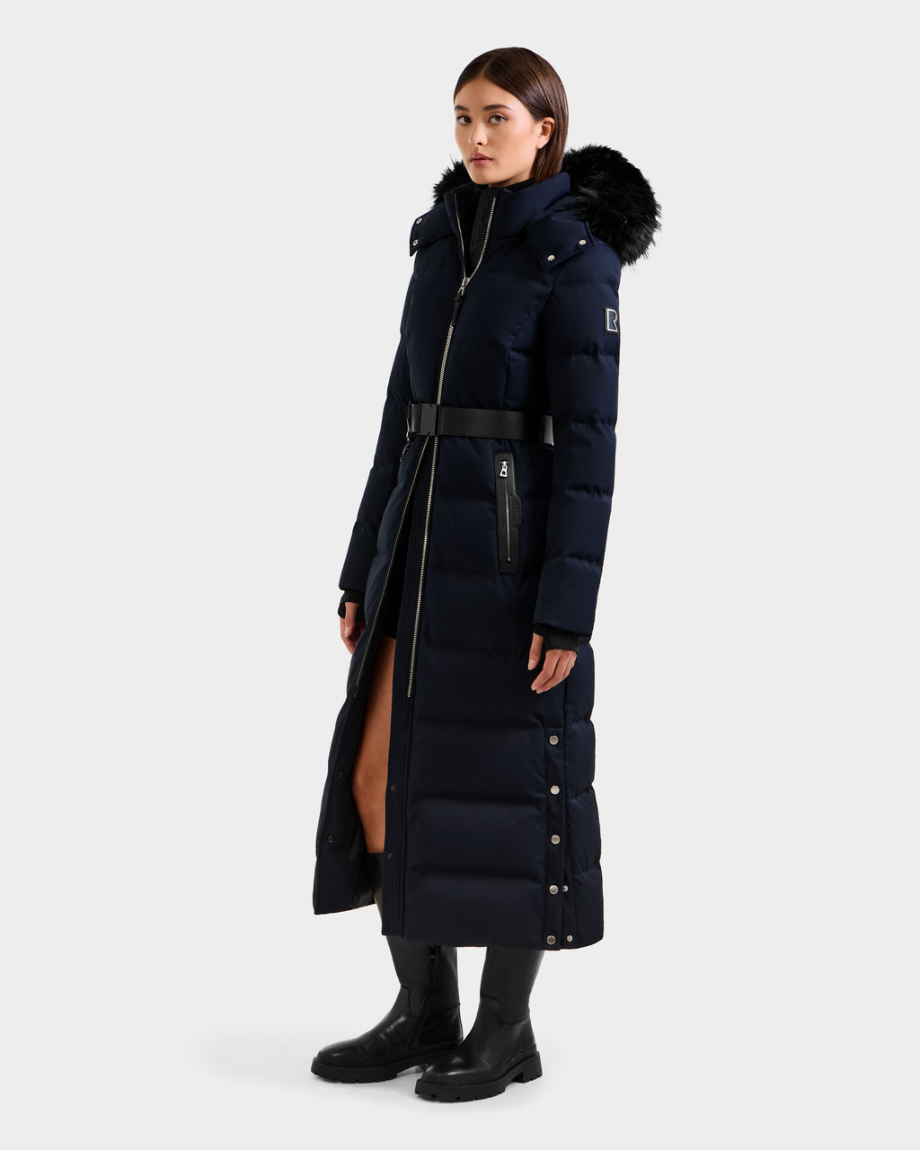 A woman wears the Rudsak IZABELLA H - 8125644 Navy, a long RDS-certified down puffer with a fur-trimmed hood, belted waist, high collar, front zipper, side snaps, and black boots against a plain background.