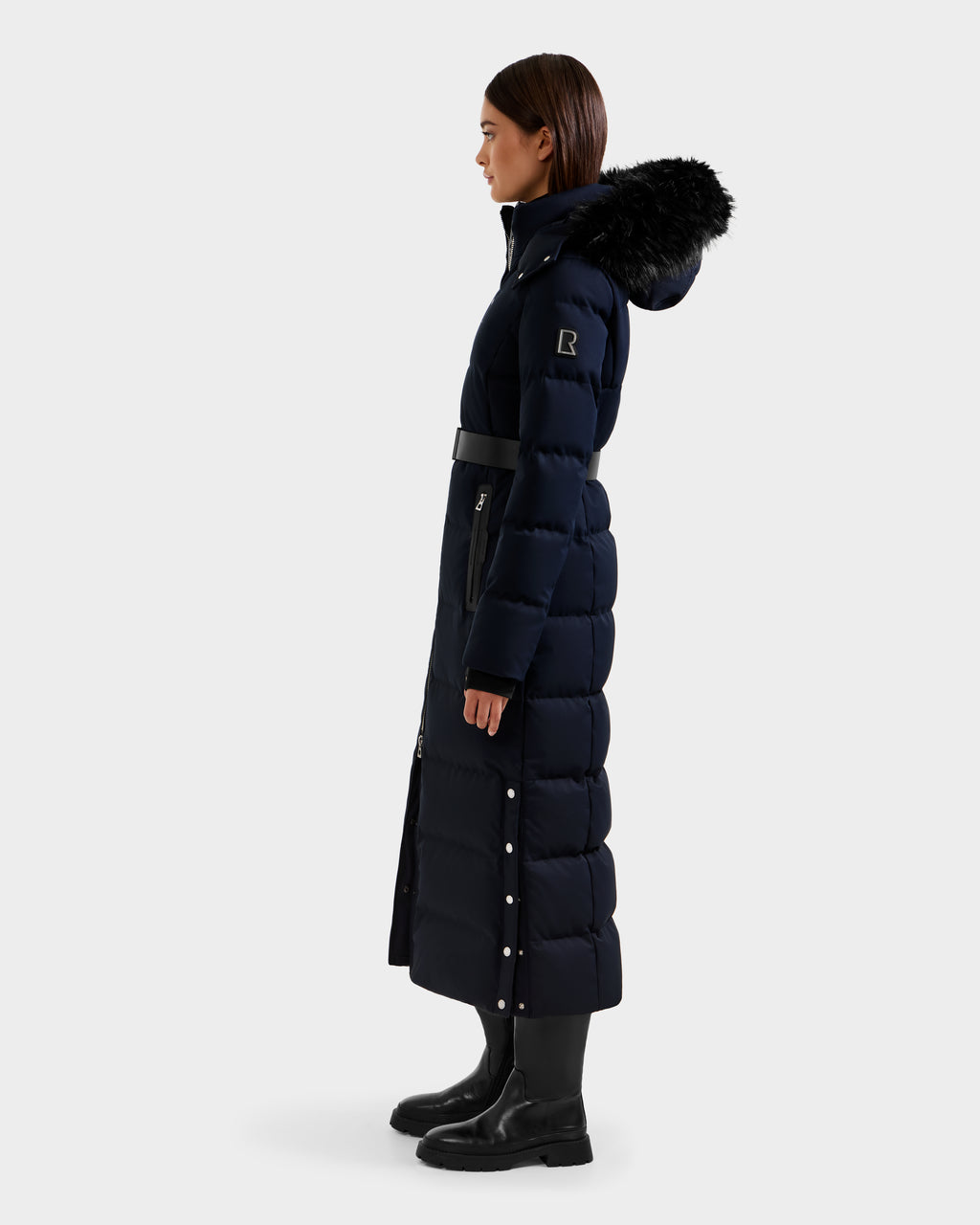 A woman in the Rudsak IZABELLA H - 8125644 Navy long RDS-certified down puffer coat with a belted waist and fur-trimmed hood, standing in profile against a plain background, wearing black boots—a stylish choice for any occasion.