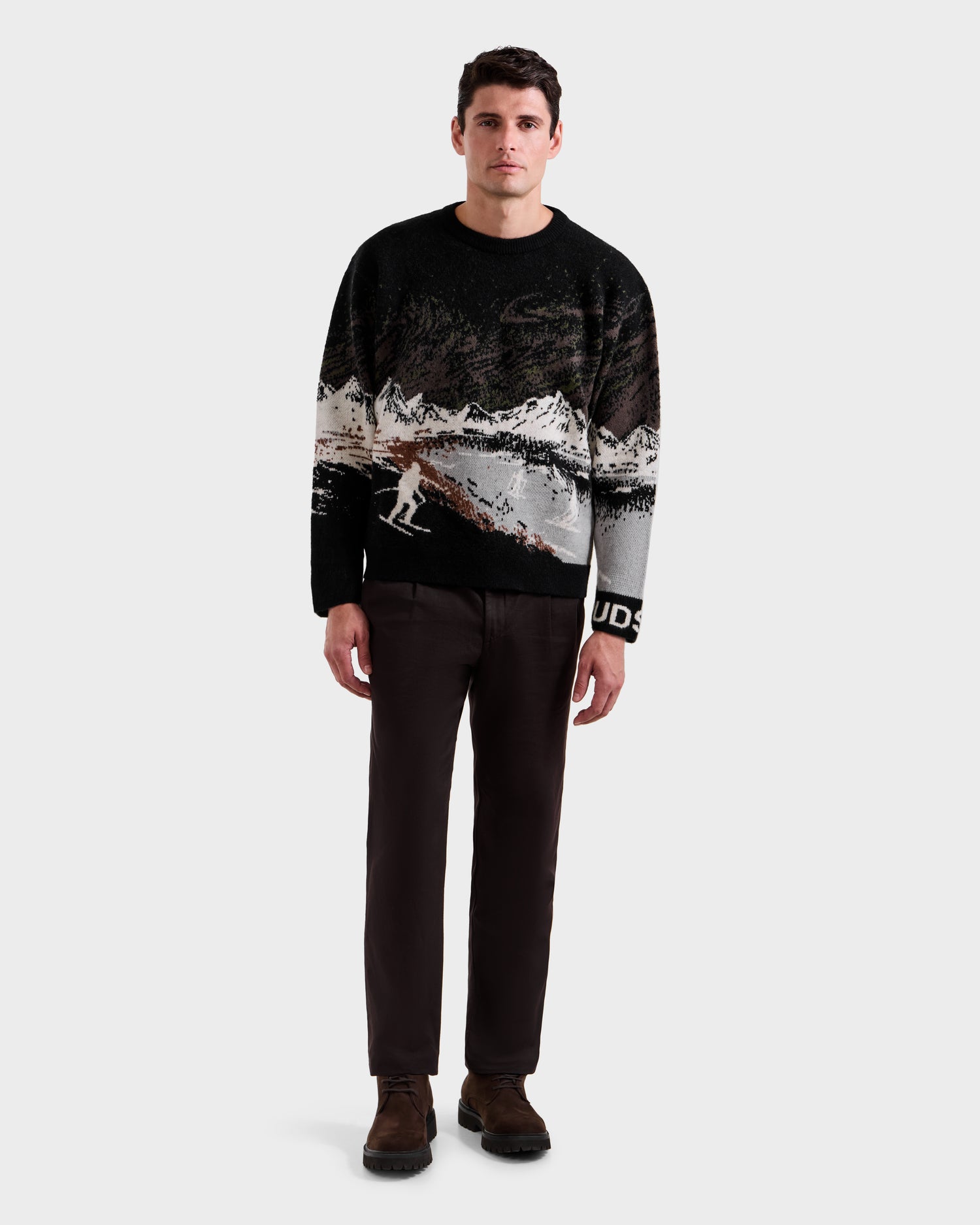A man stands facing forward in an alpine setting, wearing the Rudsak IZZY - 9525557 Black M unisex knit sweater with dark brown pants and brown shoes, set against a plain light gray background.