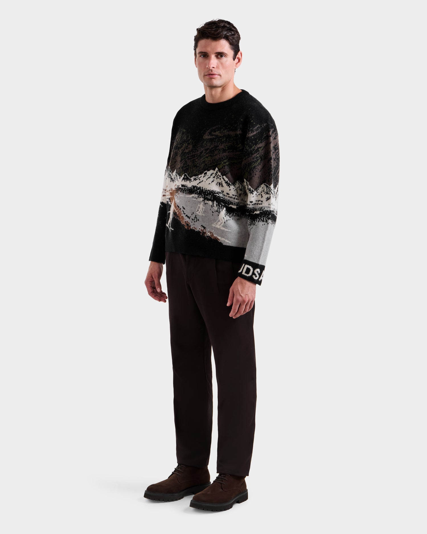 A man faces forward in the Rudsak IZZY - 9525557 Black M unisex knit sweater with a mountain landscape design, dark brown pants, and brown shoes, showcasing an alpine look against a plain white background.