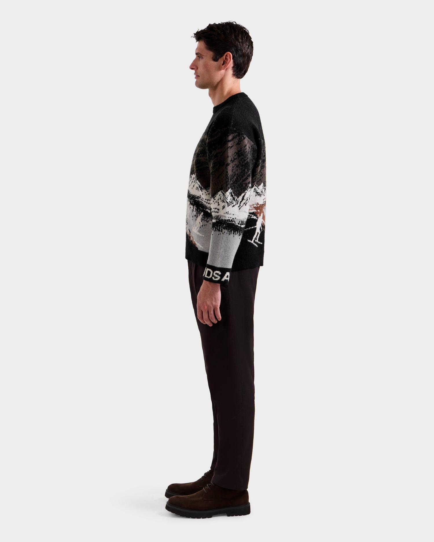 A man stands in profile against a plain background, wearing the Rudsak IZZY - 9525557 Black M sweater with a mountain landscape design, paired with dark pants and brown boots for an alpine-inspired look.