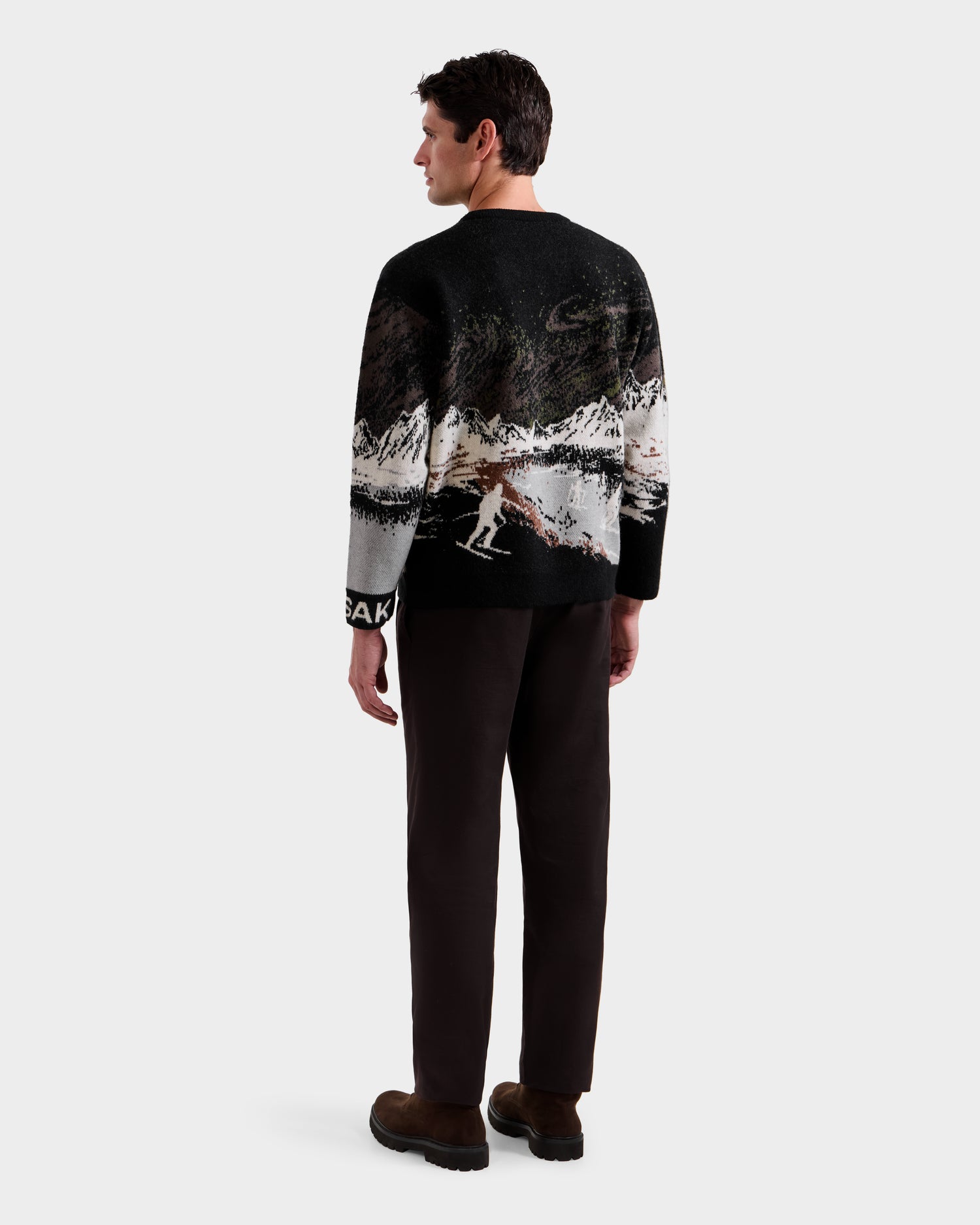 A man faces away wearing the Rudsak IZZY - 9525557 Black M unisex knit sweater with a mountain and skier design, paired with dark pants and brown shoes, set against a plain white background.