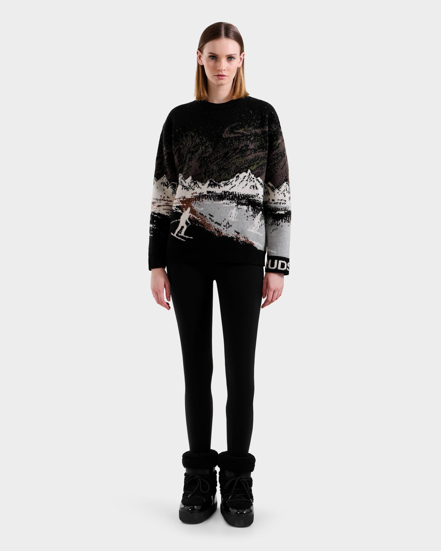 A woman stands facing forward, wearing Rudsak's IZZY - 9525557 Black W winter boots, black leggings, and a black and white alpine crewneck with a mountain and skier design against a plain light gray background.