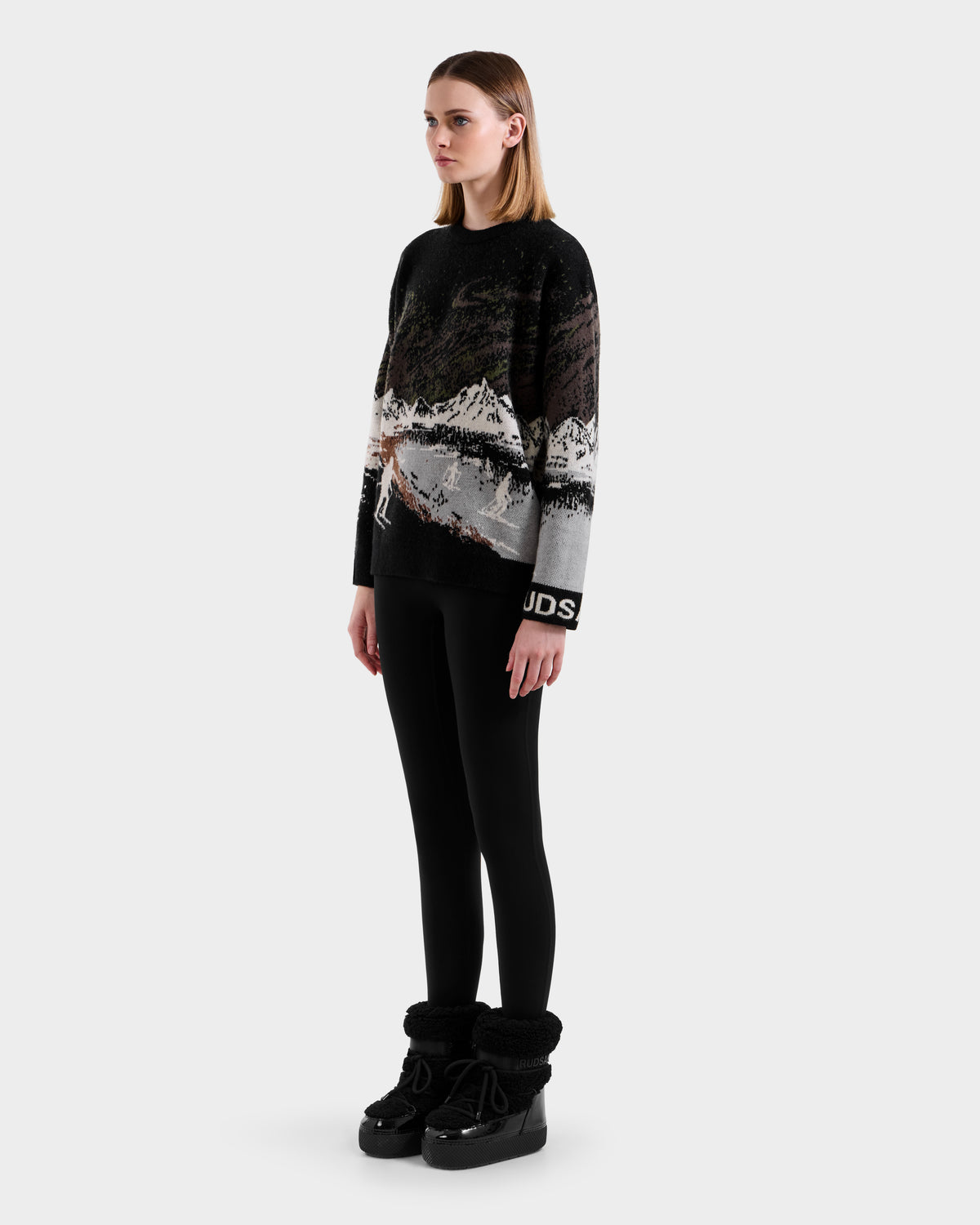 A woman with straight, shoulder-length hair and a neutral expression wears the Rudsak IZZY - 9525557 Black W alpine crewneck sweater featuring a mountain landscape, paired with black leggings and fur-trimmed boots, against a plain background.