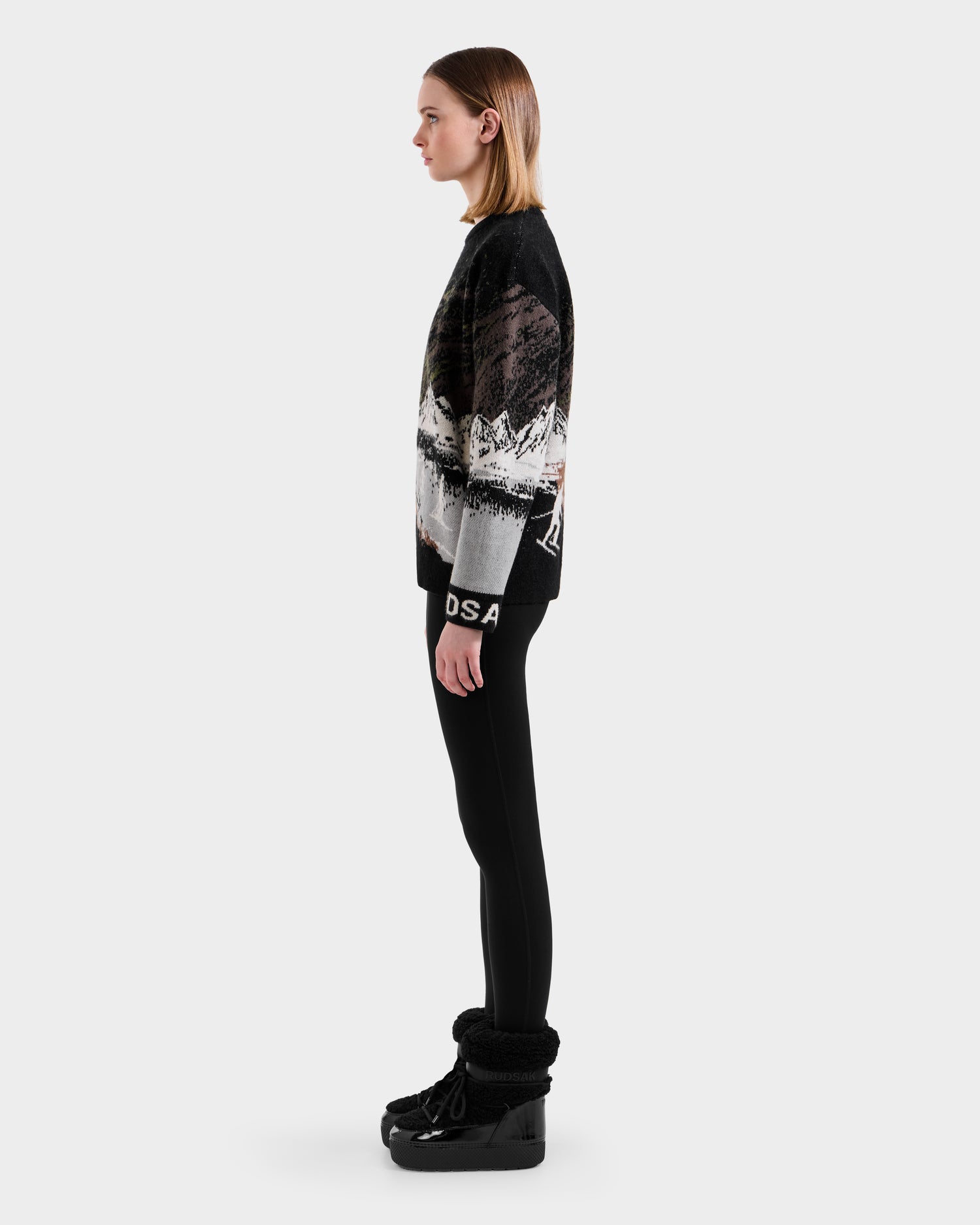 A woman stands in profile against a plain background, wearing an alpine crewneck sweater with a mountain landscape design, black DS4 leggings, and Rudsak IZZY - 9525557 Black W boots.