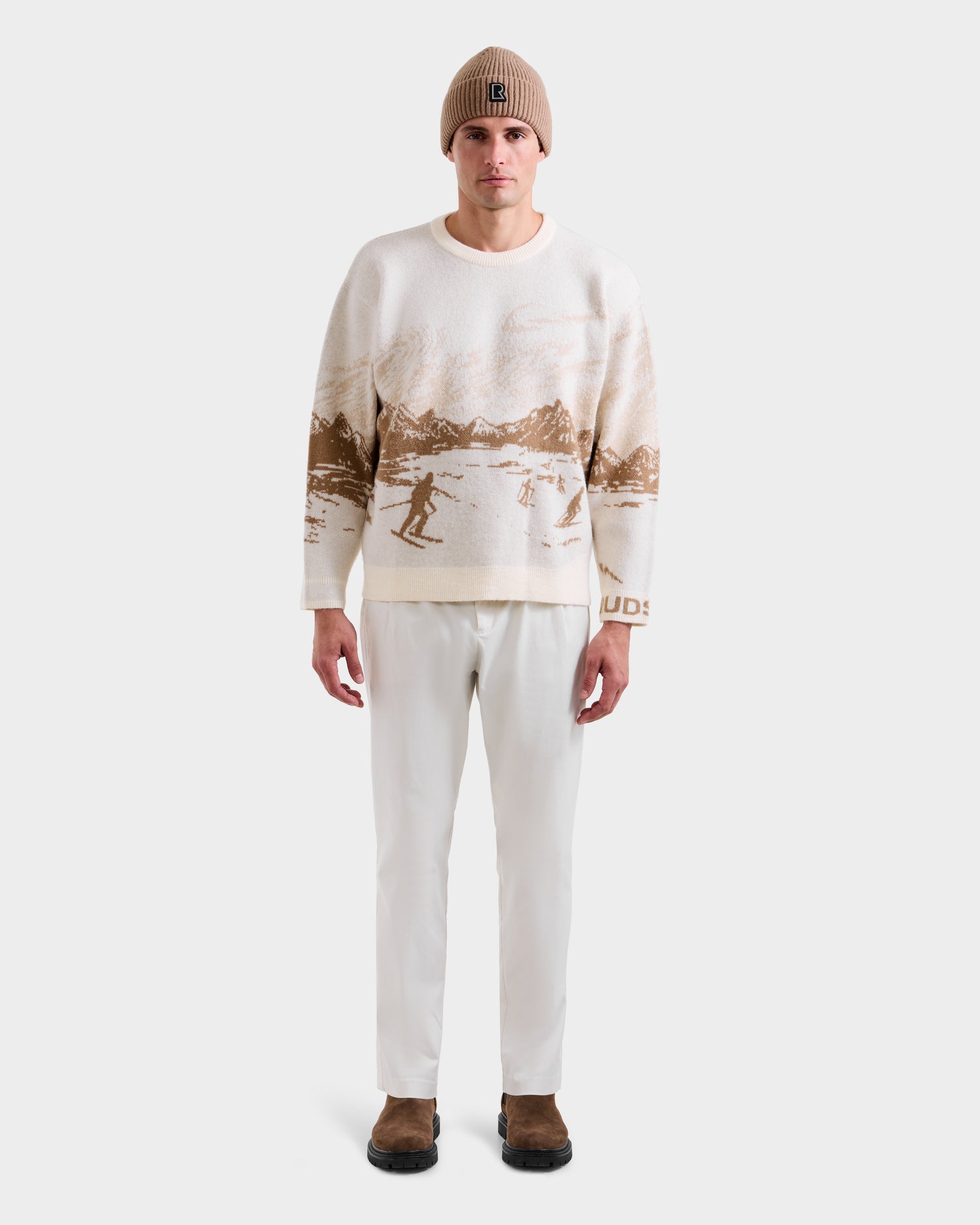 A man wearing the Rudsak IZZY - 9525557 Iceland M unisex knit crewneck sweater with a brown ski mountain motif, a beige beanie, white pants, and brown shoes stands facing forward against a plain white background.