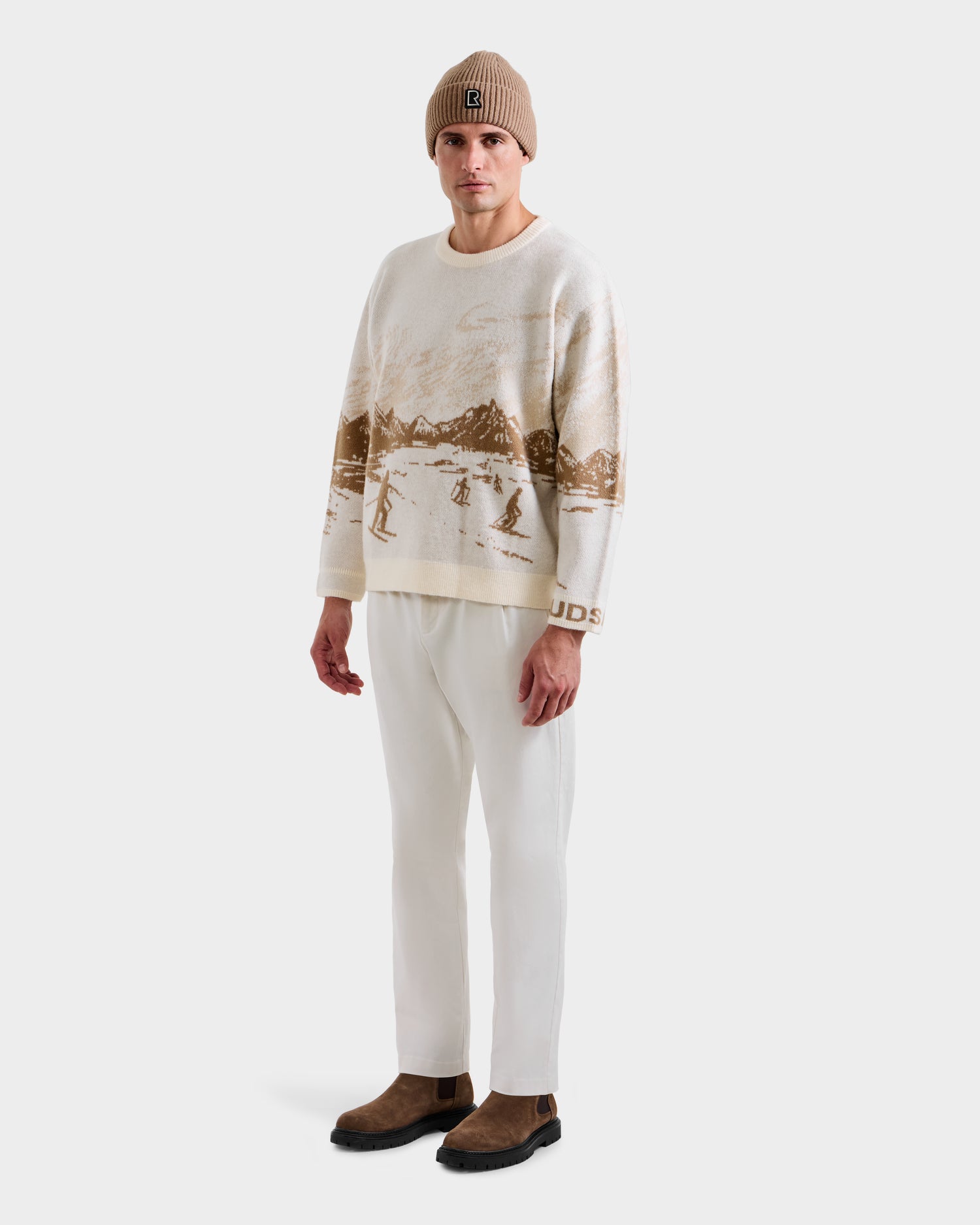 A man wearing the Rudsak IZZY - 9525557 Iceland M knit beanie, a unisex knit crewneck with a brown ski mountain motif, white pants, and brown Chelsea boots stands facing forward against a plain white background.