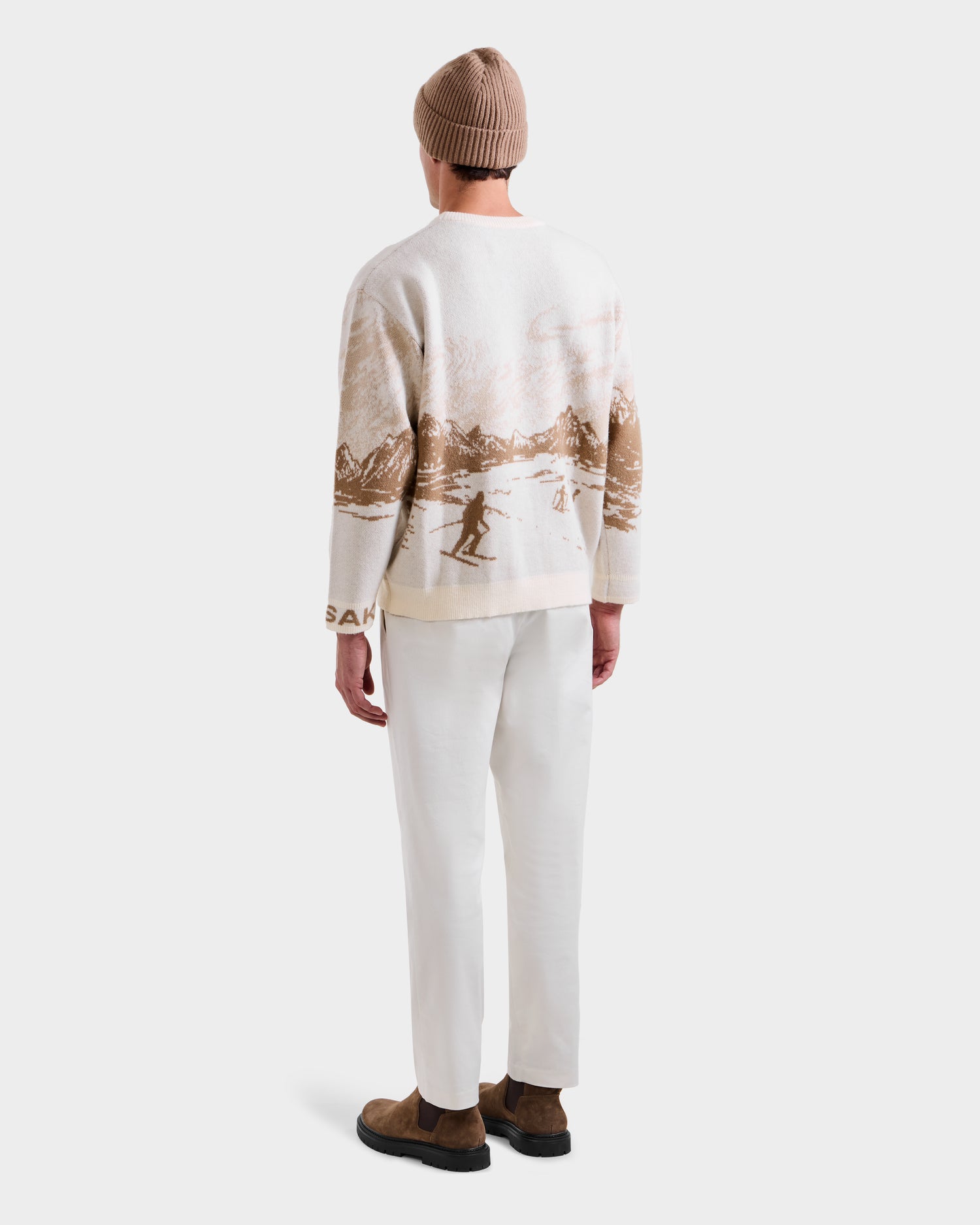A person stands facing away against a plain white background, wearing the Rudsak IZZY - 9525557 Iceland M unisex knit crewneck sweater with a ski mountain motif, paired with a light brown beanie, white pants, and brown shoes.