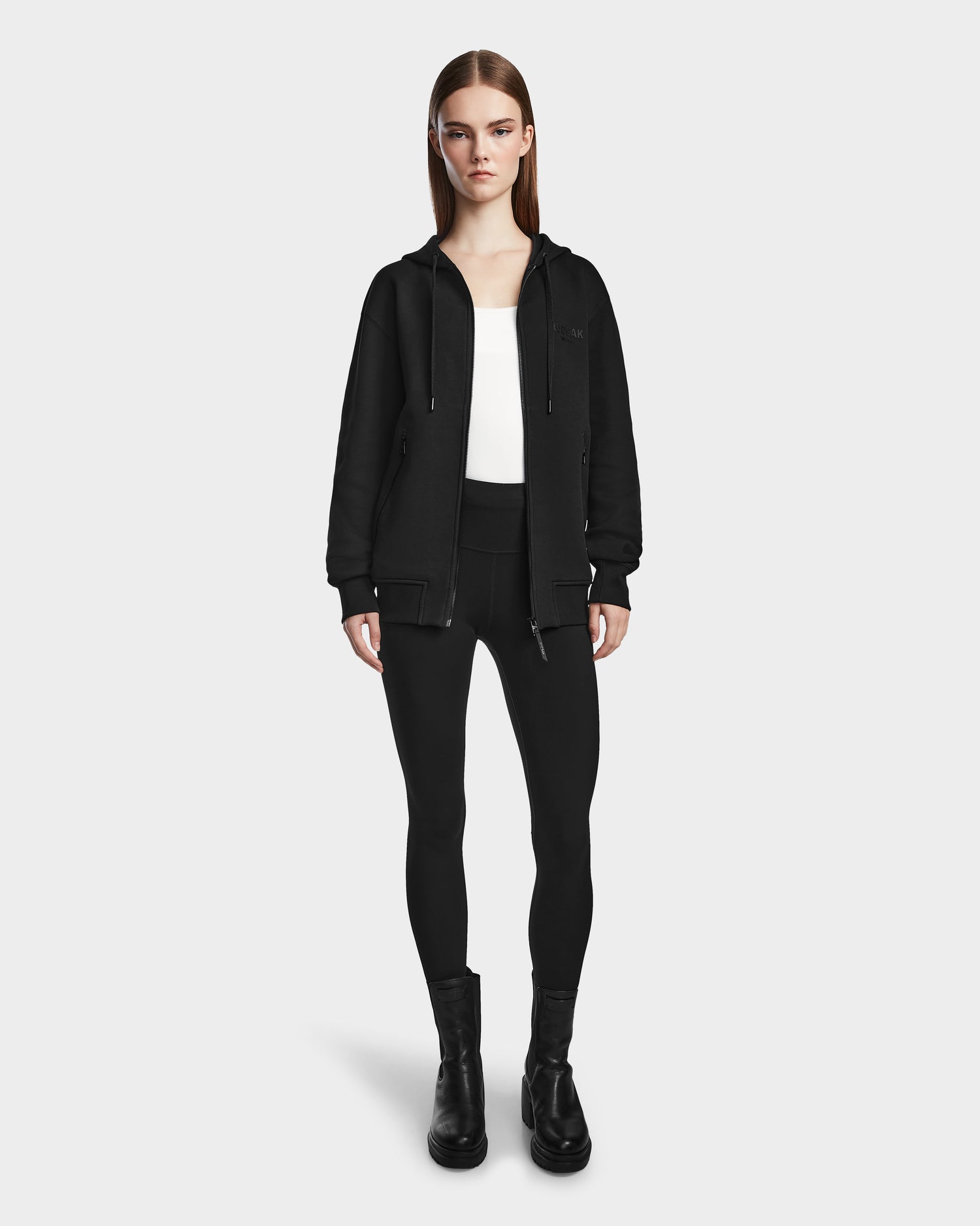 A woman stands facing forward in a Rudsak JACE - 9524010 Black W hoodie, black leggings, white top, and black ankle boots against a plain white background.