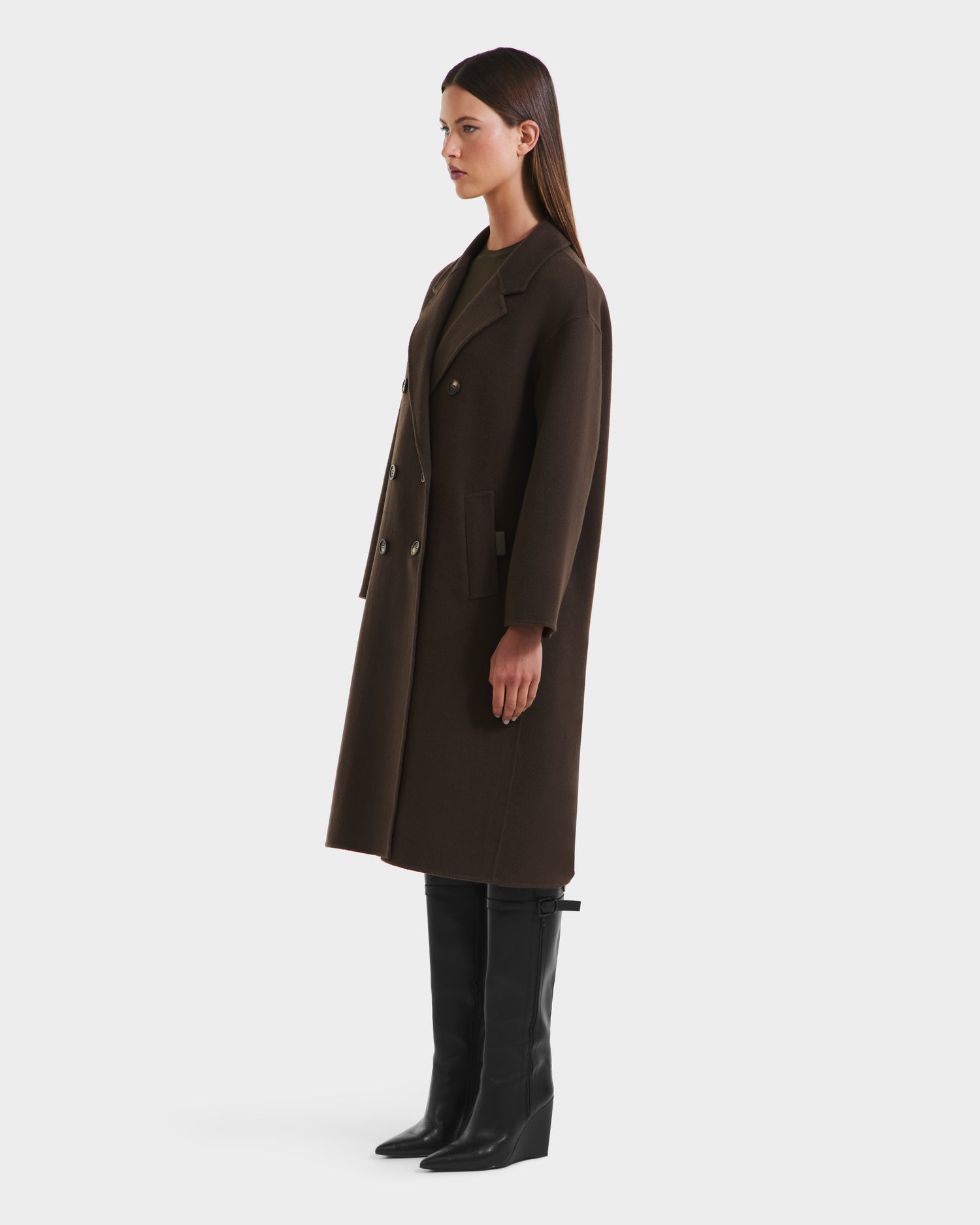 A woman in profile wears the JADE - 8125673 Army double-breasted coat by Rudsak with black knee-high boots; her straight hair is center-parted. The background is plain white.