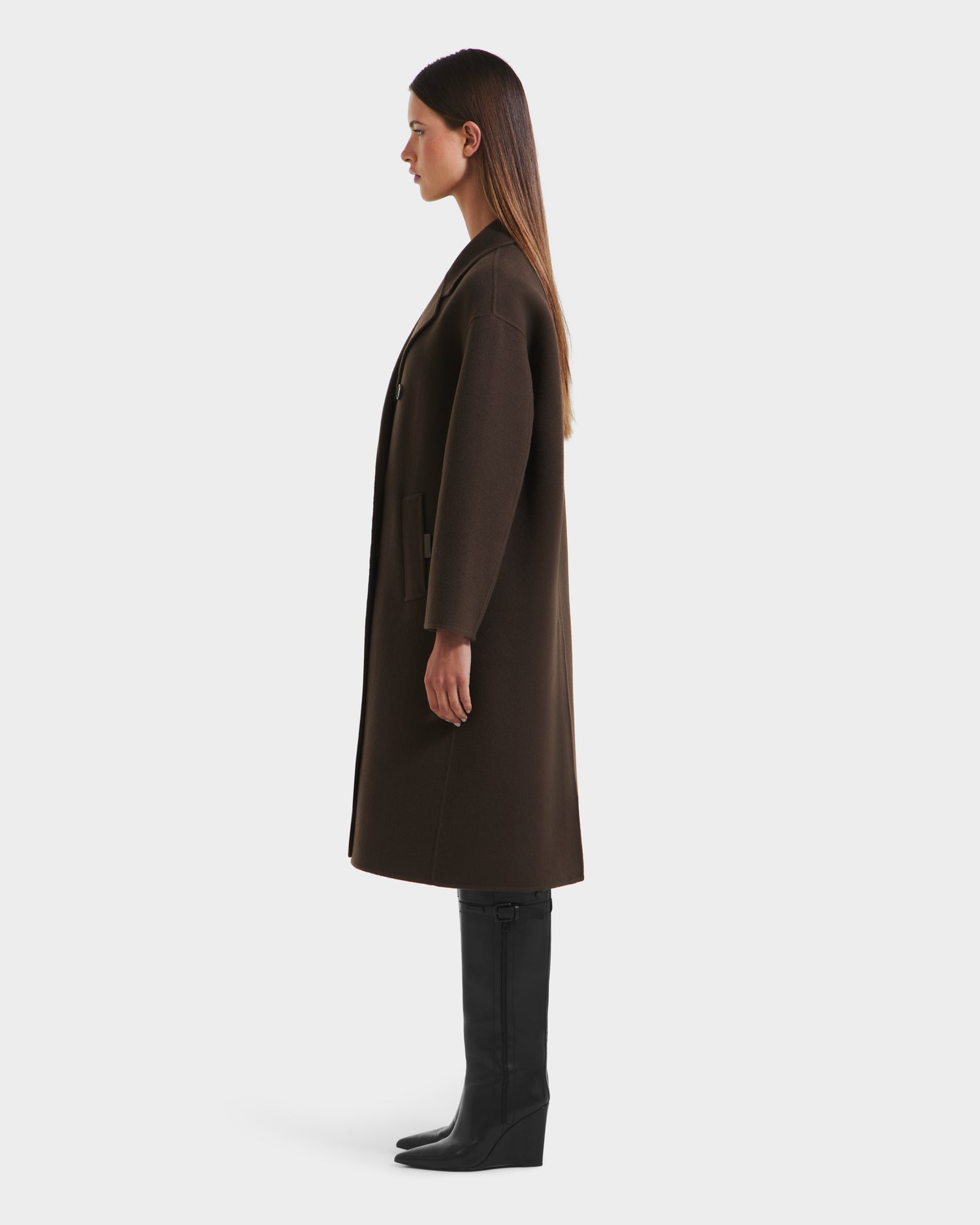 A woman stands in profile against a white background, wearing the Rudsak JADE - 8125673 Army double-breasted coat and knee-high black wedge boots. Her straight hair falls loose as she looks ahead with arms at her sides.