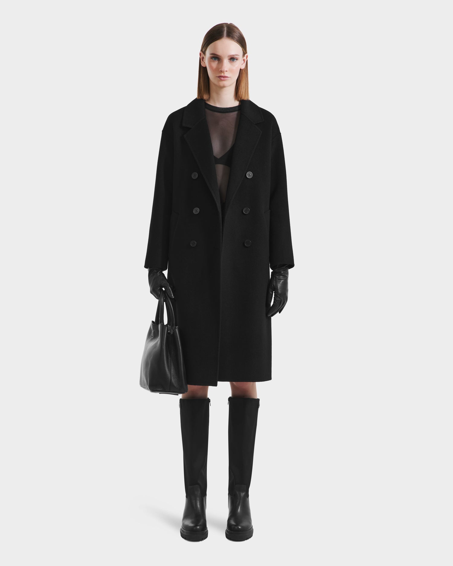 A woman faces forward wearing the Rudsak JADE - 8125673 Black double-breasted coat, paired with a sheer black top, black gloves, knee-high boots, and a black handbag against a plain light background.