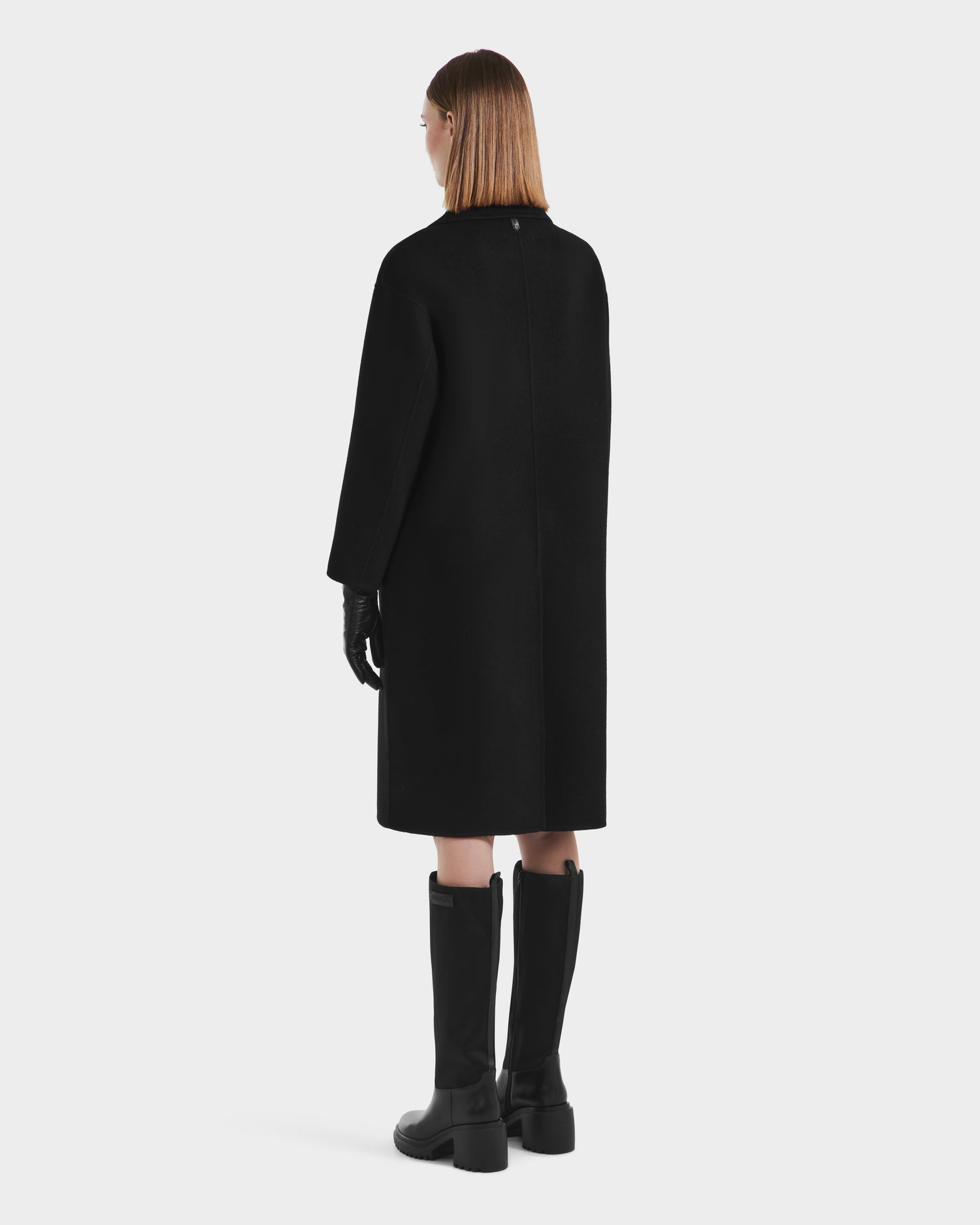 A woman with straight, light brown hair stands facing away in the Rudsak JADE - 8125673 Black women’s wool overcoat, long black gloves, and knee-high black boots against a plain white background.
