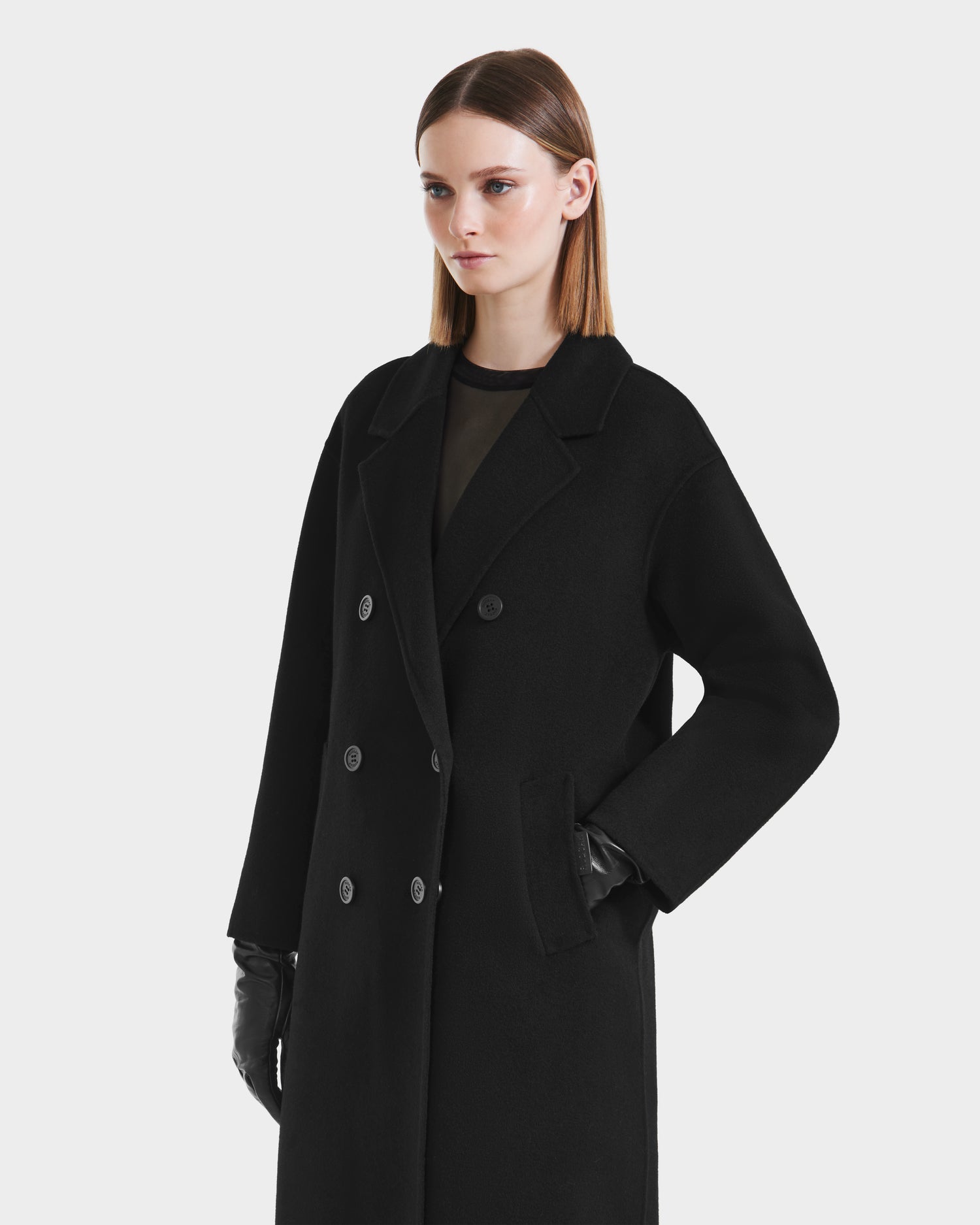 A woman with straight, shoulder-length brown hair models the Rudsak JADE - 8125673 Black double-breasted wool-blend coat and black gloves, standing with one hand in her pocket against a plain white background.
