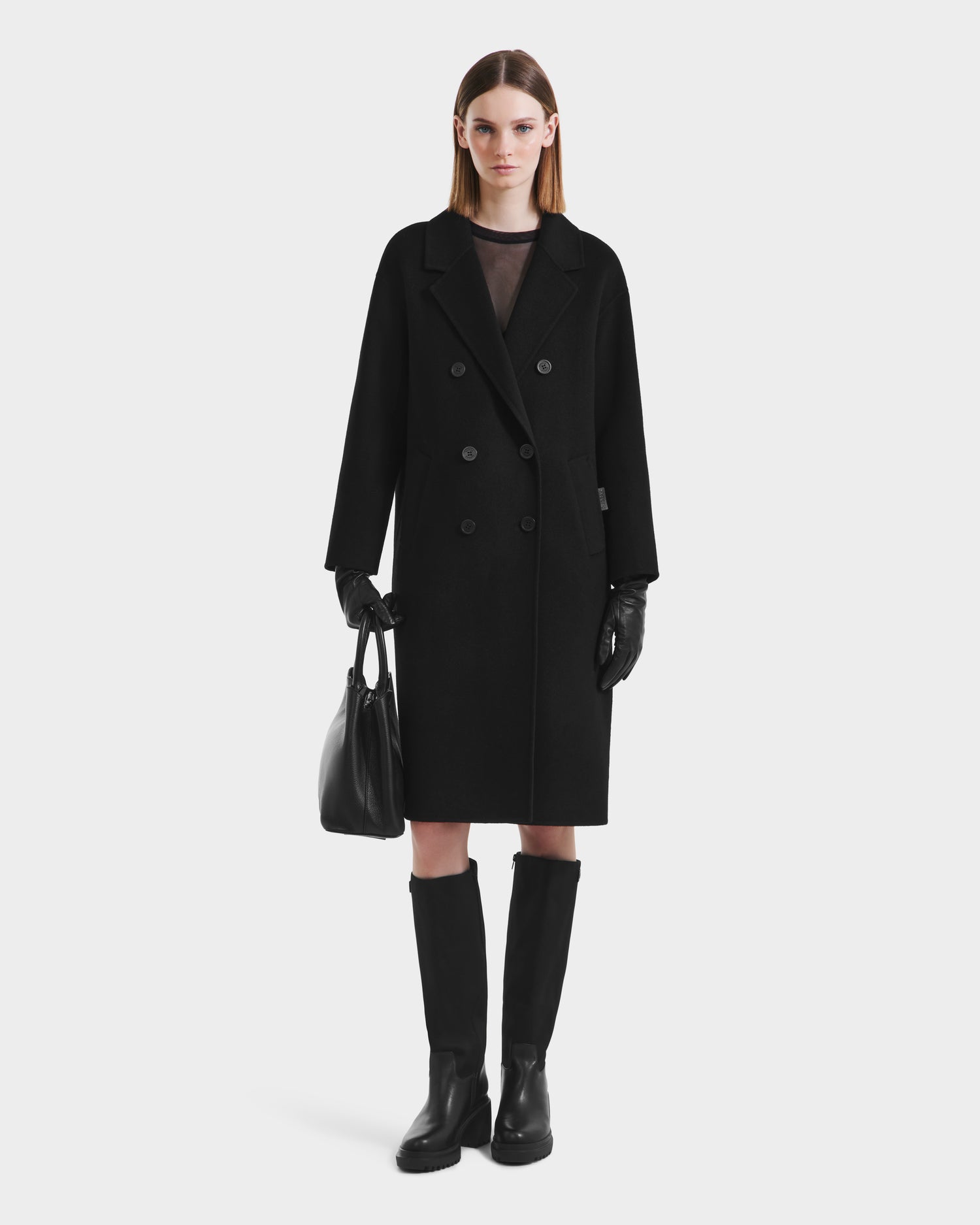 A woman stands facing forward in the Rudsak JADE - 8125673 Black coat, paired with black gloves, knee-high boots, and a sleek handbag. She has straight, shoulder-length hair and a neutral expression against a plain background.