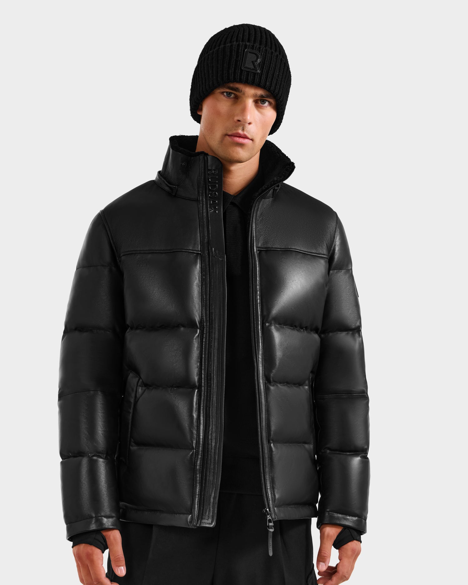 A man in a black beanie and the Rudsak JAMESON - 6125678 Black luxury men's puffer jacket stands facing forward against a plain light gray background.