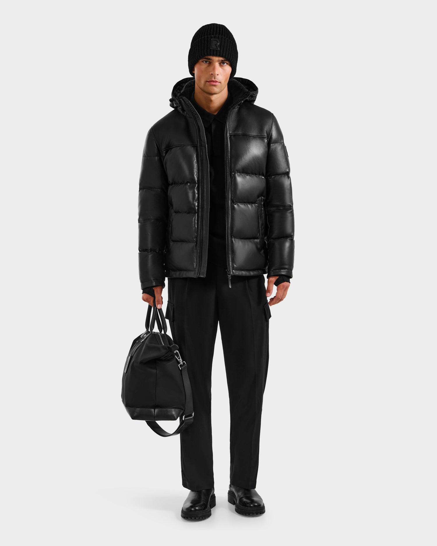 A person stands against a plain white background wearing the Rudsak JAMESON - 6125678 Black quilted leather down jacket, black pants, knit hat, shoes, and holding a black duffle bag in their right hand.