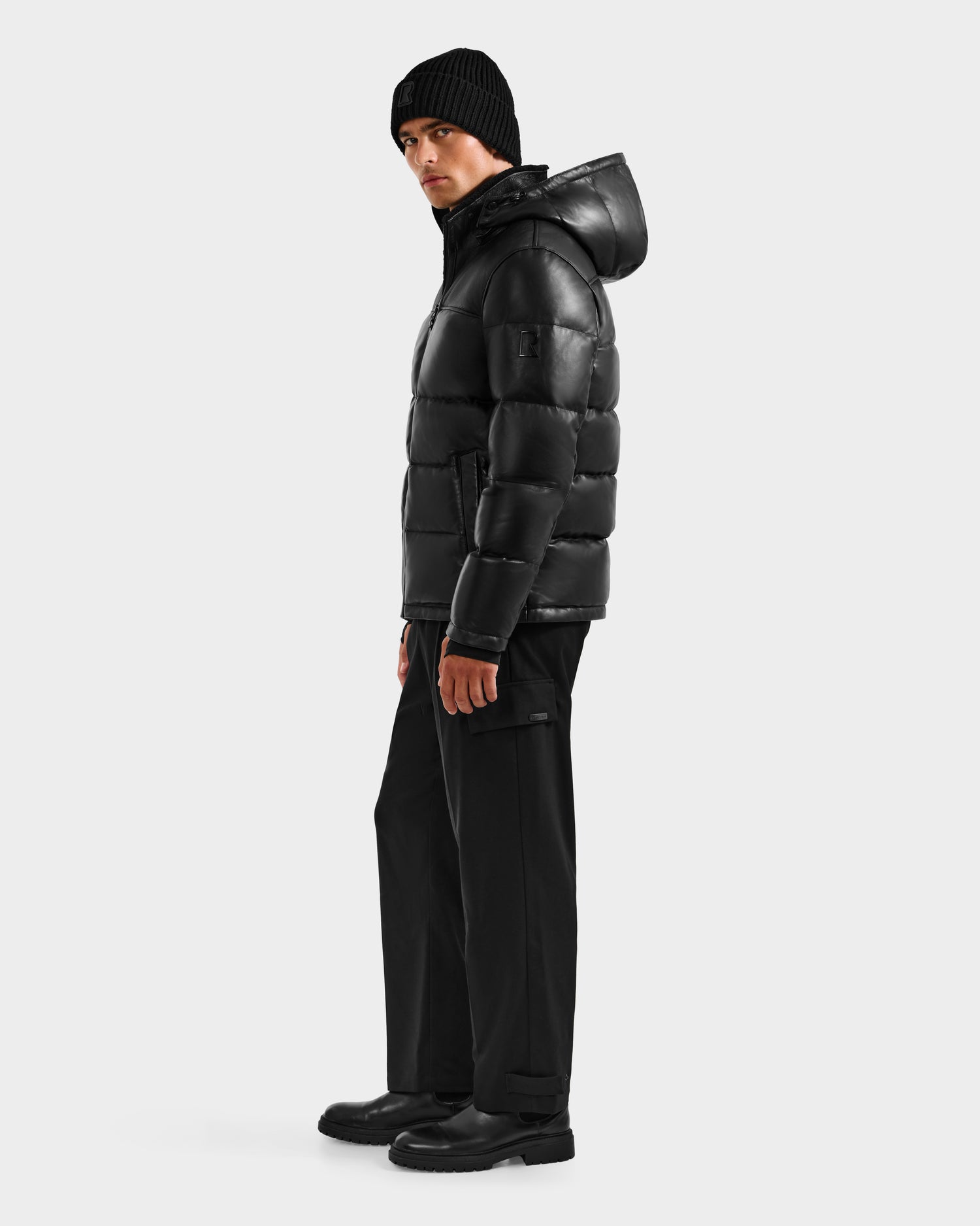 A man stands sideways against a plain light background wearing the Rudsak JAMESON - 6125678 Black men's winter jacket with hood, paired with a black beanie, black pants, and black boots.