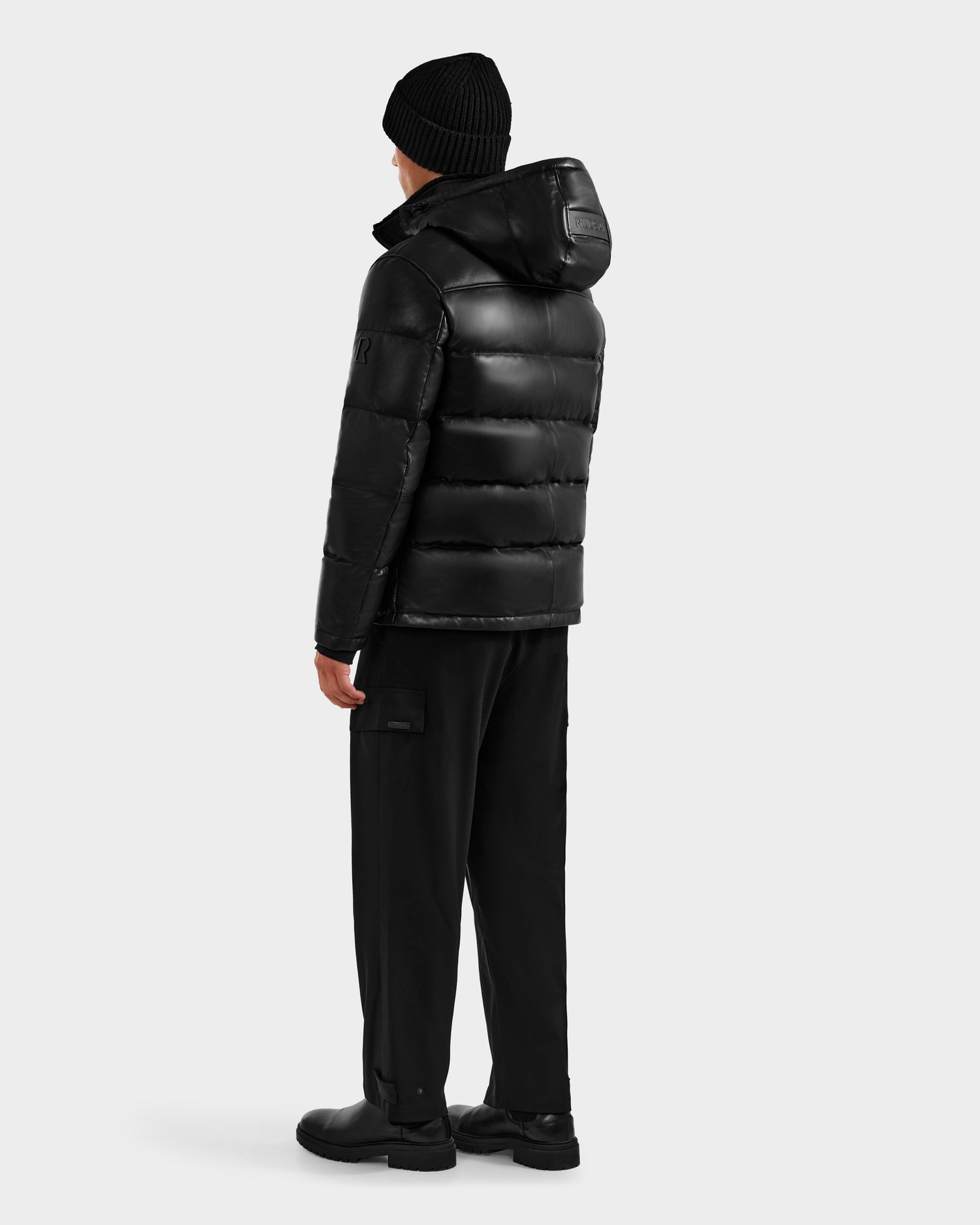 A person wearing the JAMESON - 6125678 Black quilted leather down jacket with hood by Rudsak, paired with a black beanie, pants, and shoes, stands facing away from the camera against a plain light gray background.