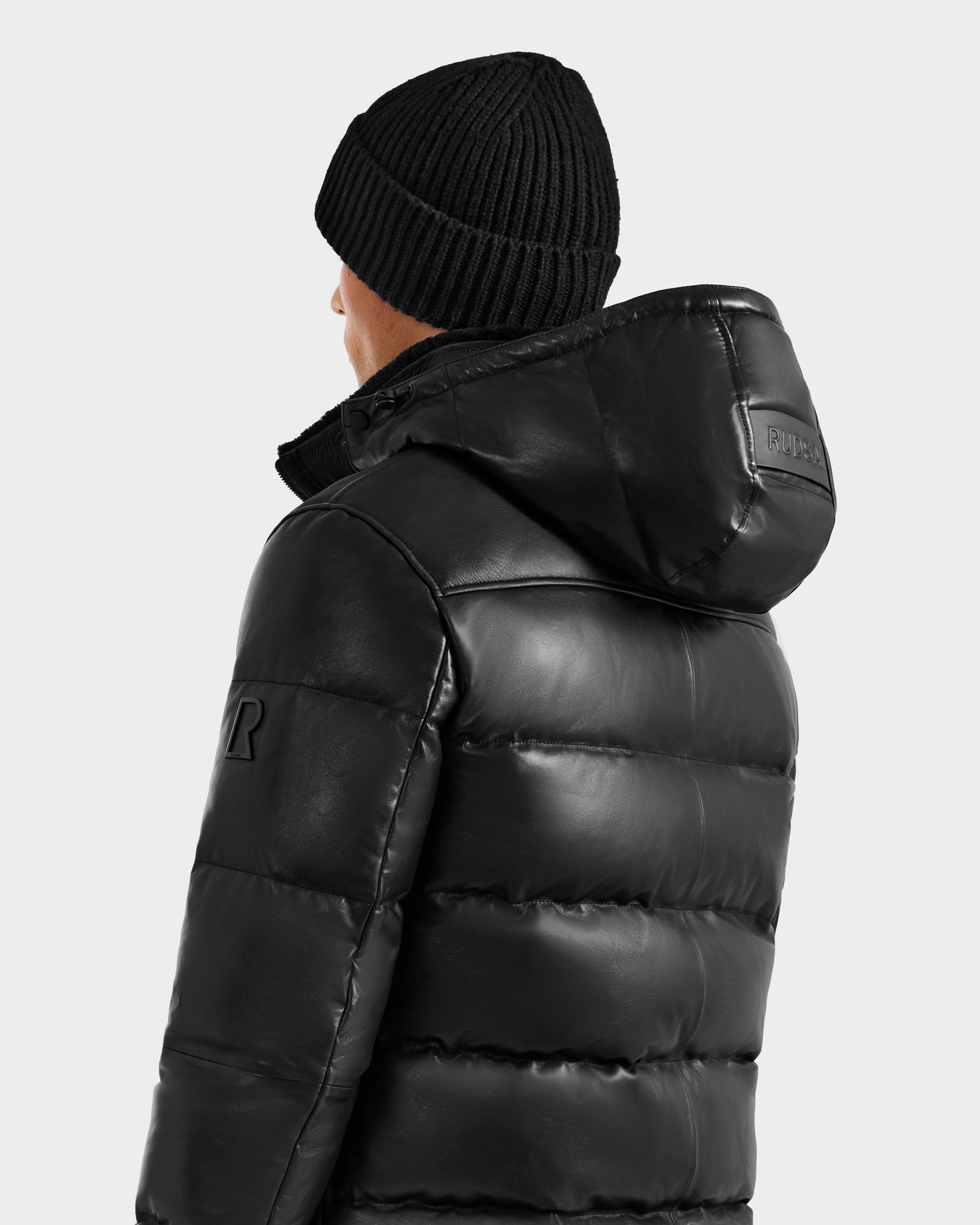 A person is pictured from the back and side wearing the Rudsak JAMESON - 6125678 Black men’s winter jacket and a black knit beanie against a plain light background.