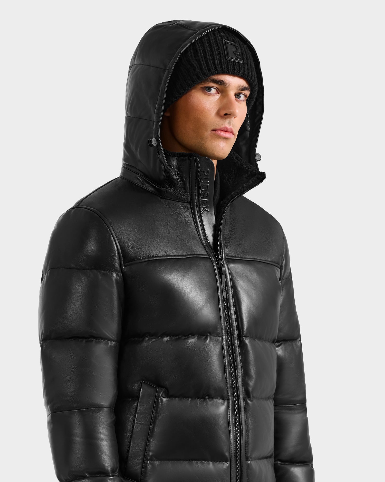 A man wears the Rudsak JAMESON - 6125678 Black puffer jacket with a hood and a black knit beanie, looking slightly to the side against a light gray background.