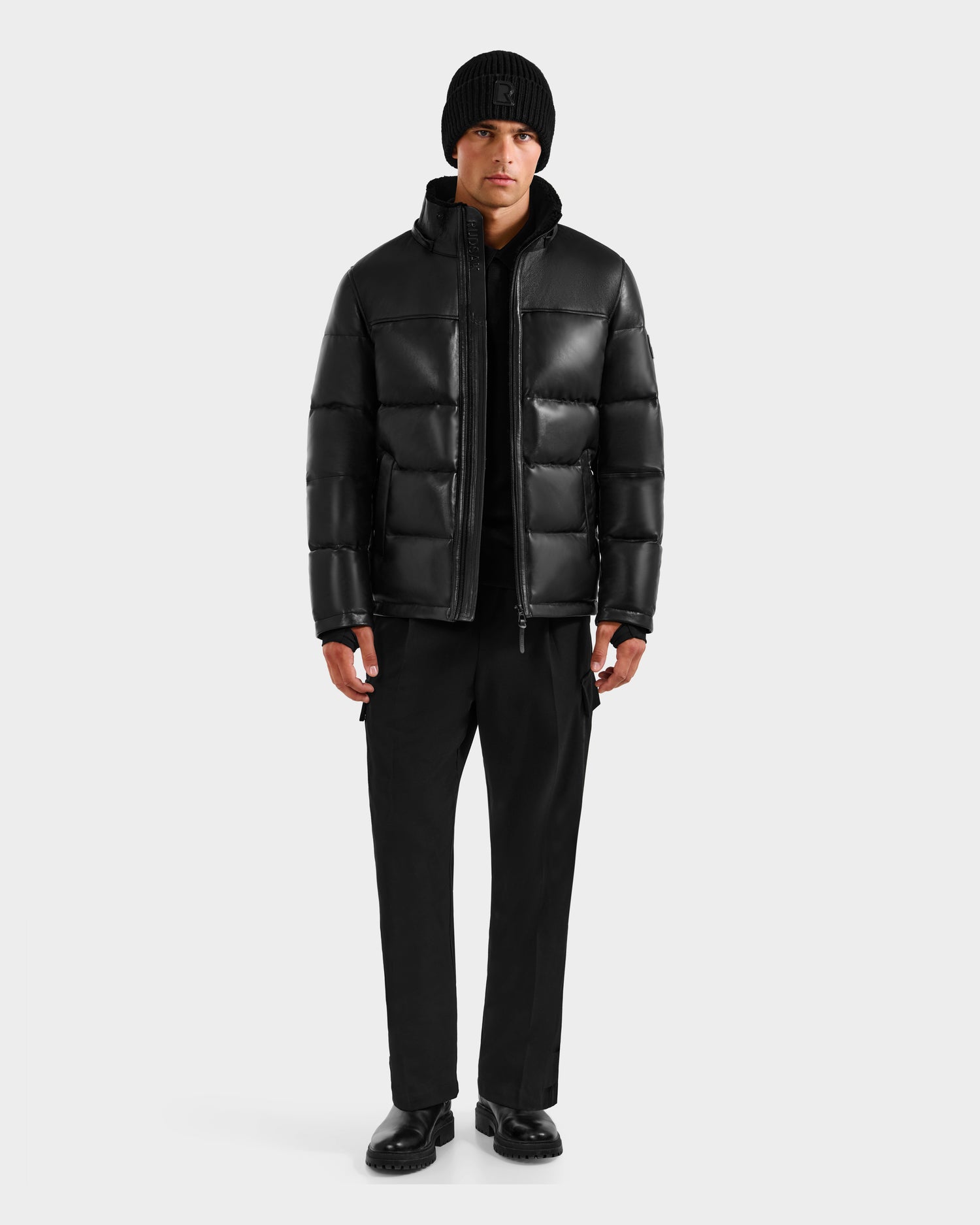 A man stands against a plain background wearing the Rudsak JAMESON - 6125678 Black puffer jacket, paired with a black knit beanie, black pants, and black shoes, facing forward with arms relaxed at his sides.