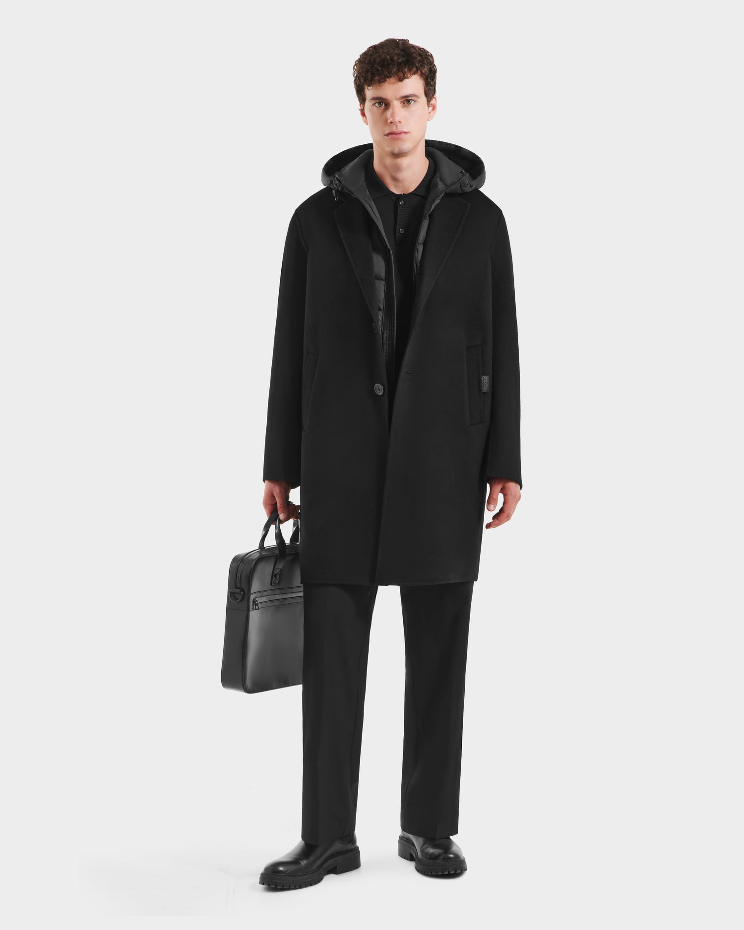 A man faces forward, wearing the Rudsak JEREMIAH - 6125509 Black men’s wool-blend coat with removable down dickie, over black pants and shoes, holding a black briefcase. The background is plain white.