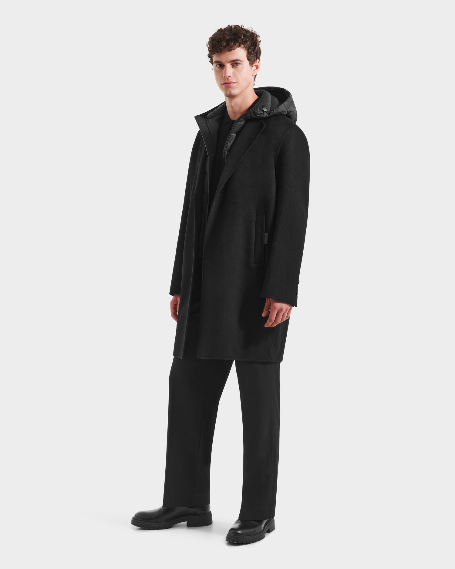 A young man with curly hair wears the Rudsak JEREMIAH - 6125509 Black men’s coat over a hoodie, black pants, and black shoes, standing against a plain white background.