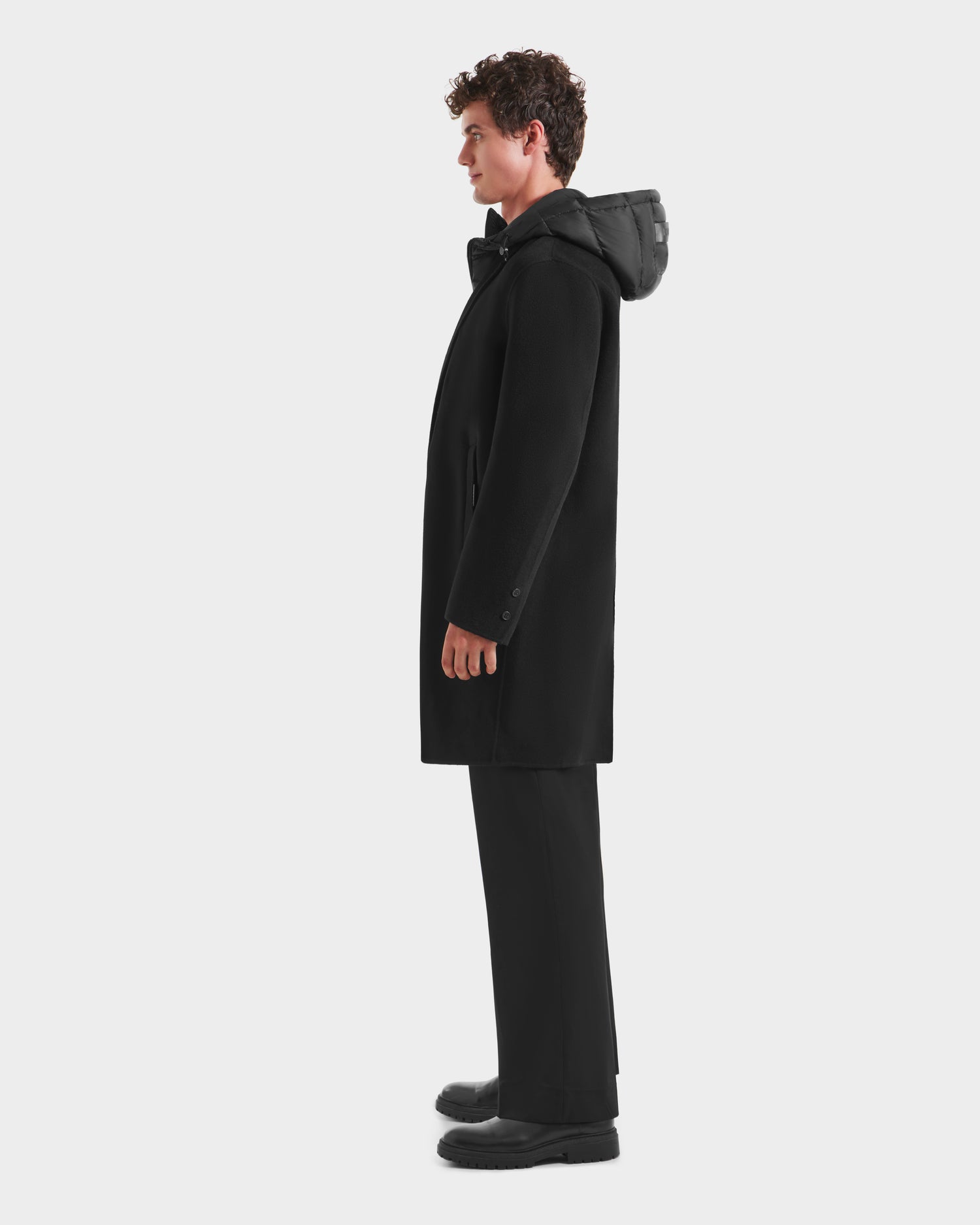 A man stands in profile against a white background, wearing Rudsak’s JEREMIAH - 6125509 Black wool-blend coat with black pants and shoes. The coat includes a removable down-insulated inner dickie for added warmth.
