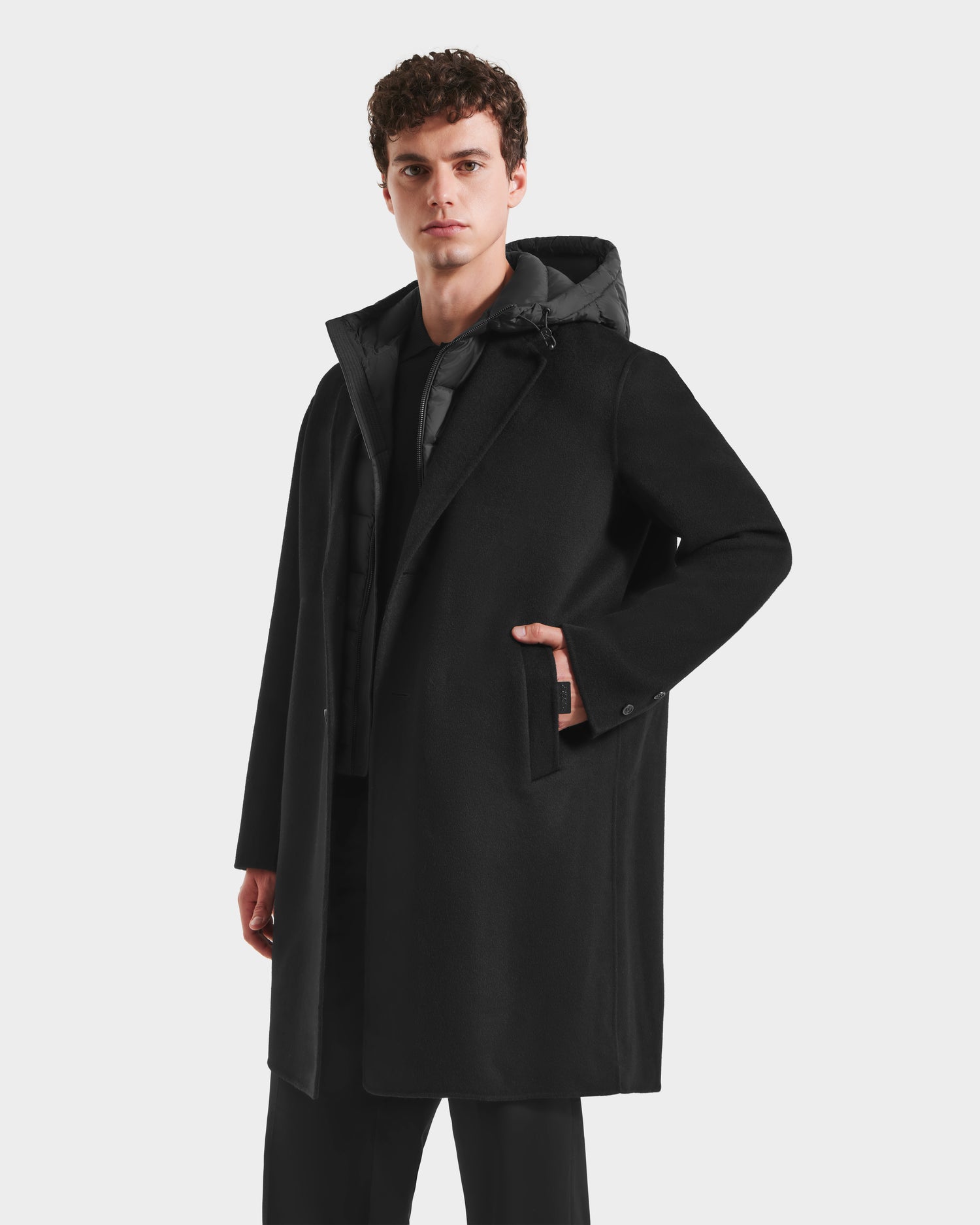 A young man with short curly hair models the Rudsak JEREMIAH - 6125509 Black men’s wool-blend coat over a dark outfit, standing with one hand in his pocket against a plain light background—a true cold-weather staple.