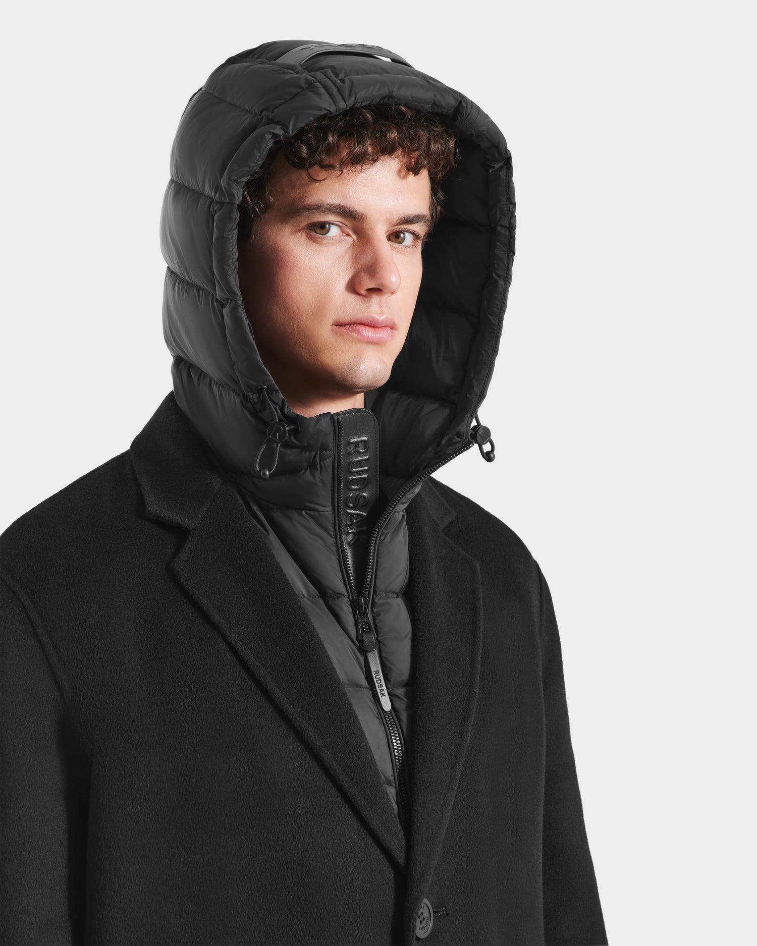 A young man with curly hair wears the Rudsak JEREMIAH - 6125509 Black hooded puffer jacket with a removable down-insulated inner dickie under a black coat, looking slightly to the side against a plain light background.