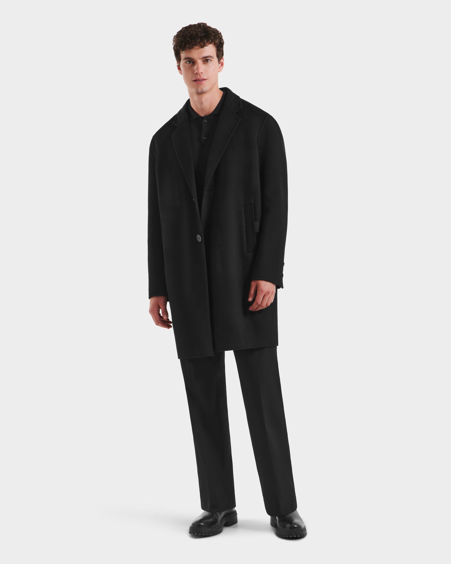 A man stands against a plain light background wearing the Rudsak JEREMIAH - 6125509 Black men’s wool-blend coat, styled with a black shirt, dress pants, and shoes. The overcoat includes a removable down-insulated inner dickie for warmth.