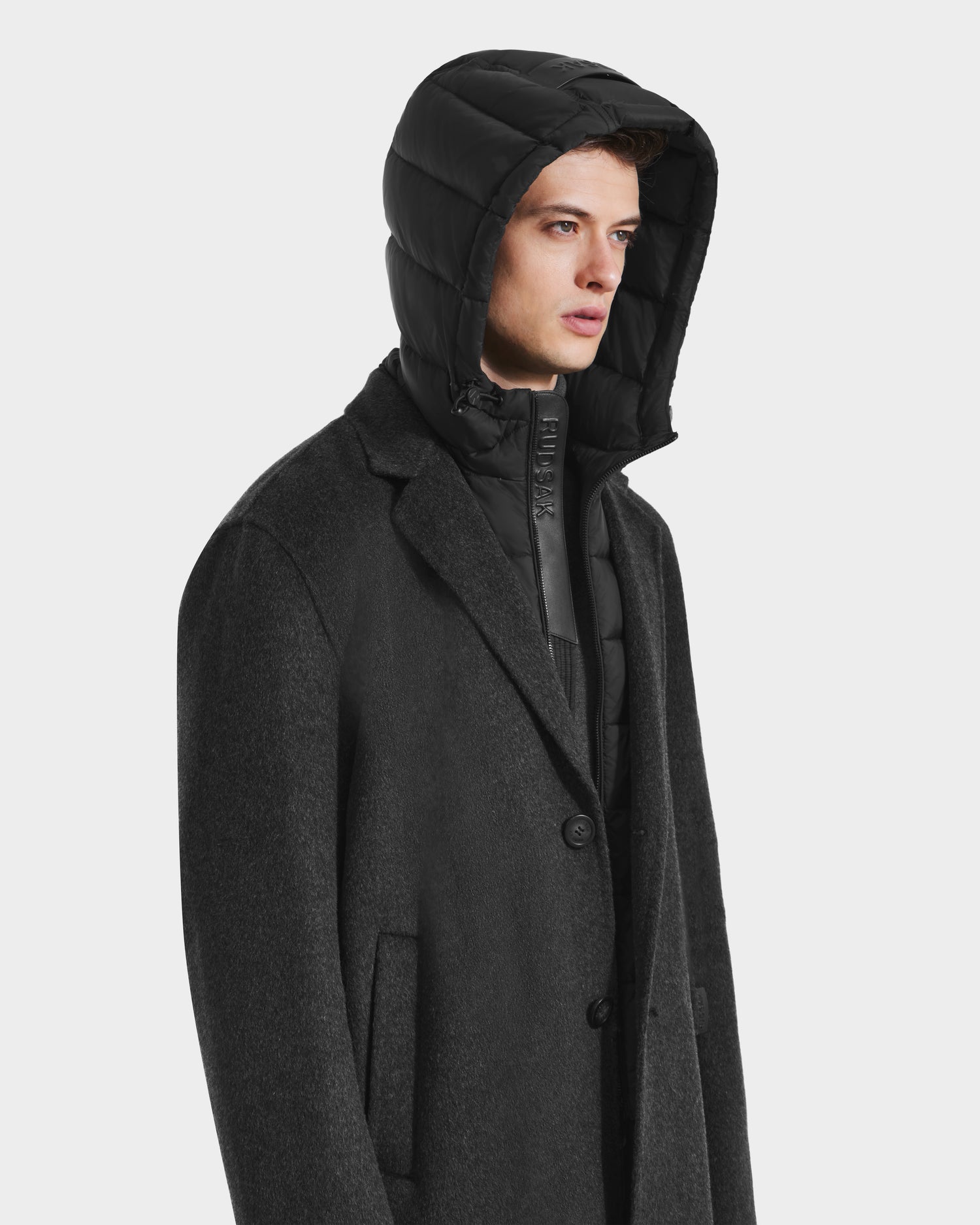 A young man in the Rudsak JEREMIAH - 6125509 Grey wool-blend coat layered over a black hooded puffer, looking to the side against a light background—a modern update on a cold-weather classic.