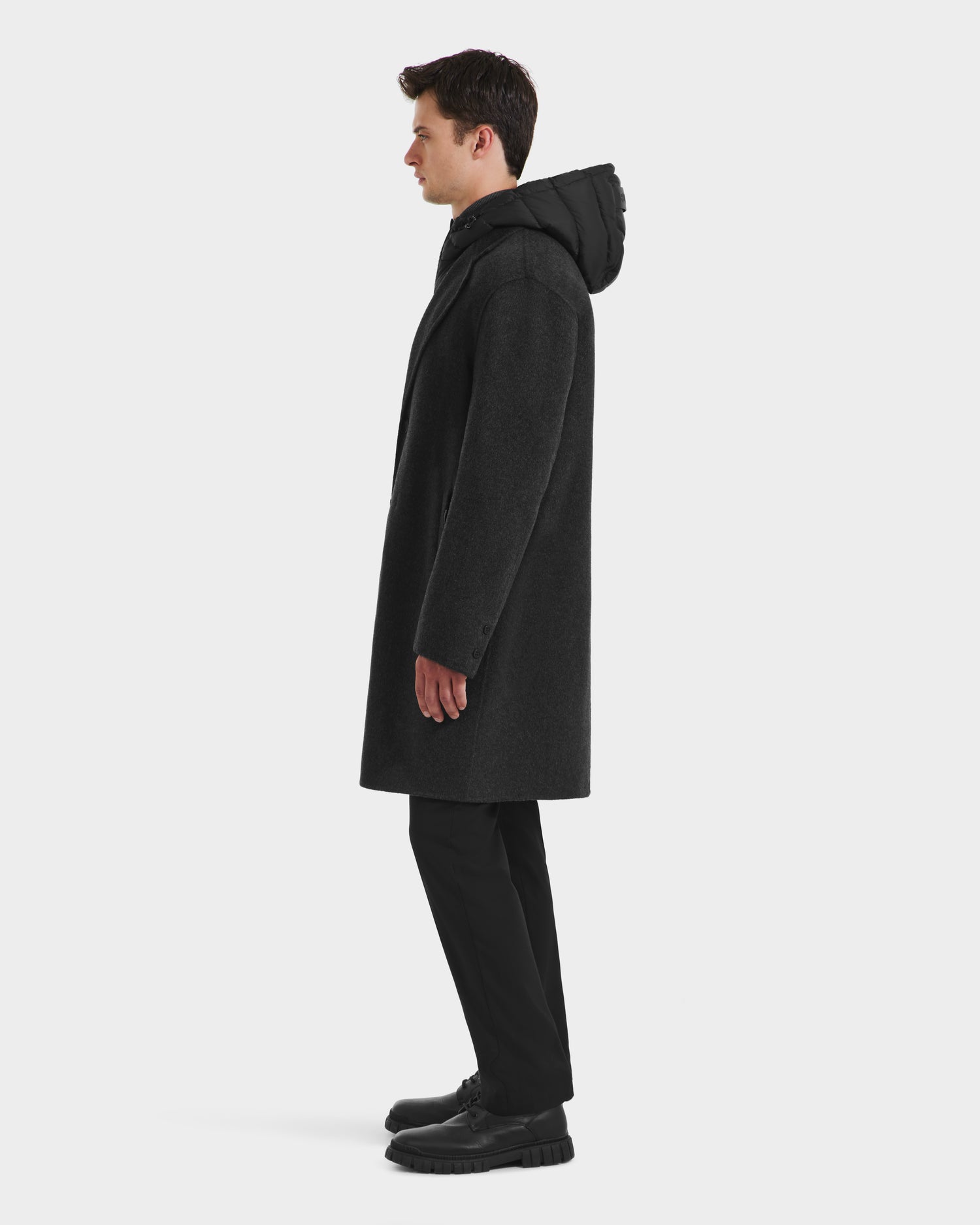 A man in profile wears the Rudsak JEREMIAH - 6125509 Grey men’s wool-blend coat with hood, paired with black pants and shoes, against a plain white background.