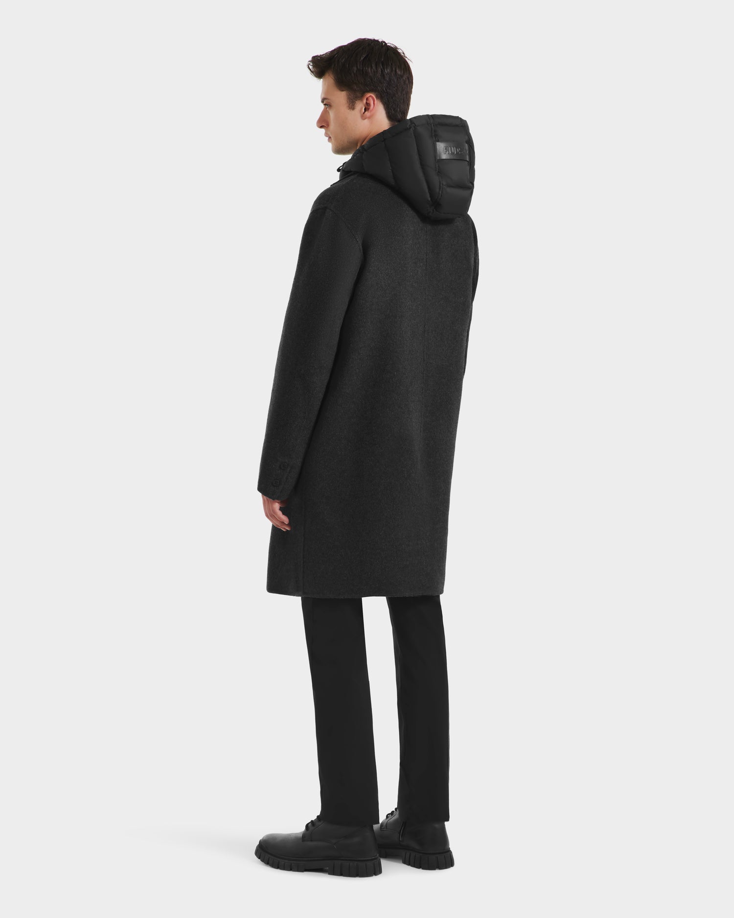 A man faces away, wearing the Rudsak JEREMIAH - 6125509 Grey men’s wool-blend coat with detachable hood and removable down dickie, paired with black pants and shoes, set against a plain white background.