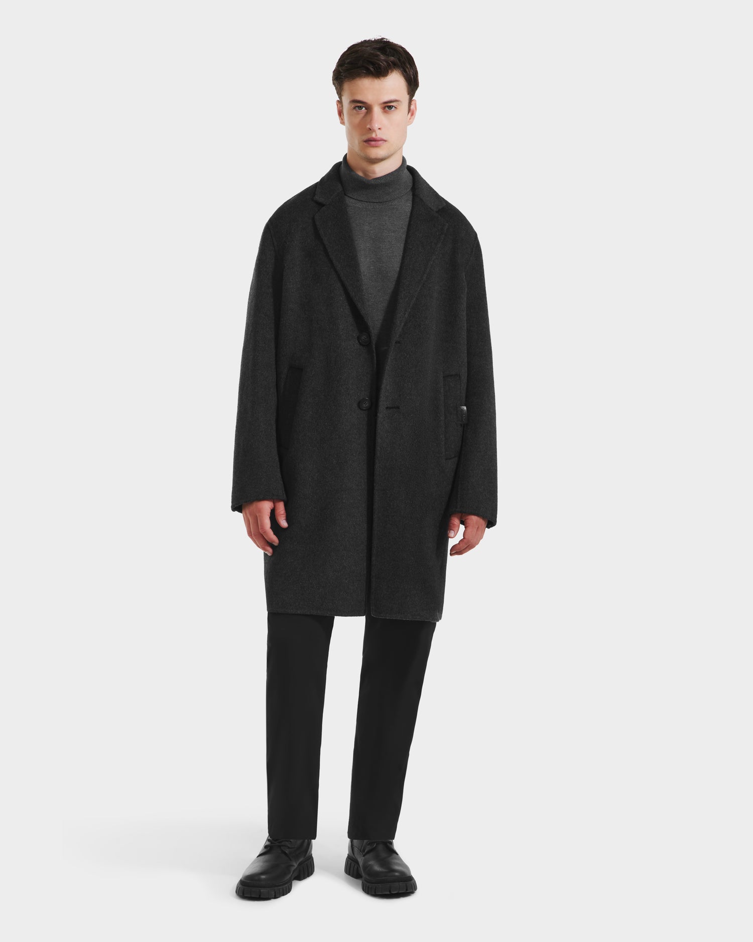 A man stands facing forward against a plain white background, wearing the Rudsak JEREMIAH - 6125509 Grey men’s wool-blend coat with a removable down-insulated inner dickie, paired with a gray turtleneck sweater, black pants, and black shoes.