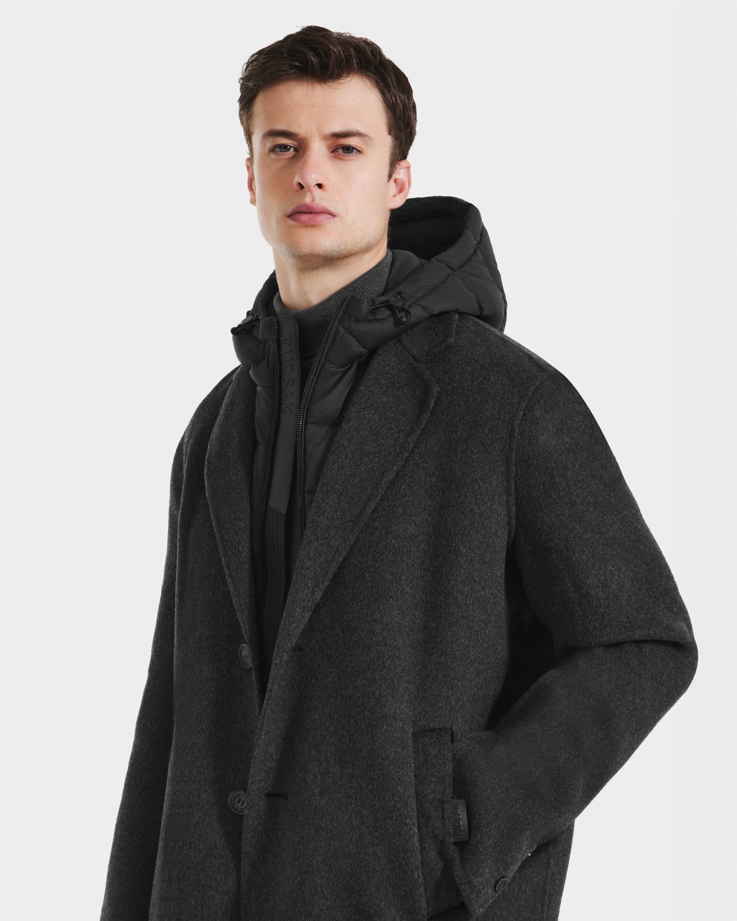 A young man with short brown hair wears the Rudsak JEREMIAH - 6125509 Grey men’s wool-blend coat with a removable down-insulated dickie over a black hooded puffer, standing against a plain light background with a neutral expression.
