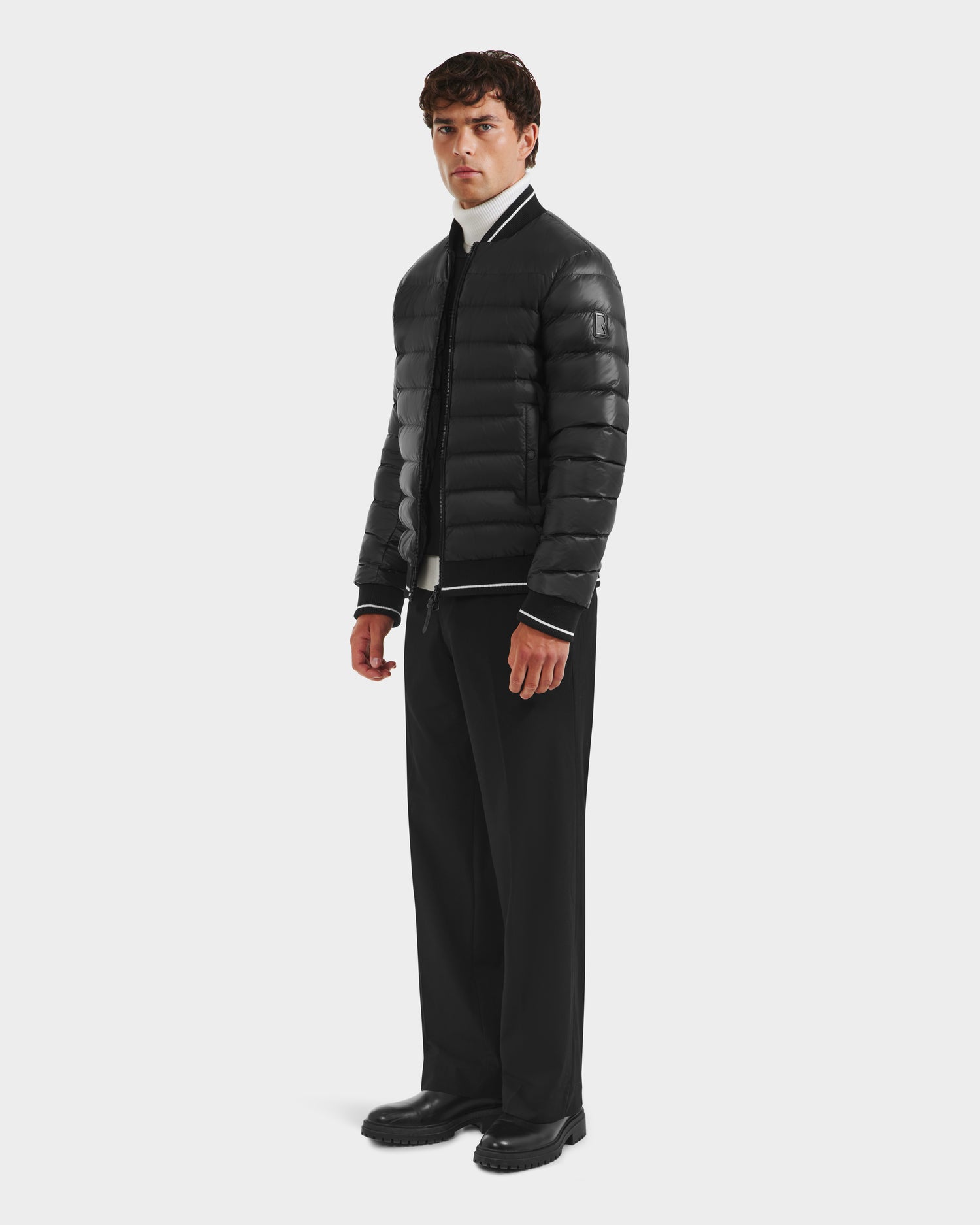 A man with short dark hair and a neutral expression stands against a white background wearing the Rudsak JEREMY - 6125025 Black men’s down bomber jacket, black wide-leg pants, a white turtleneck, and black shoes—ideal for minimalist wardrobes.