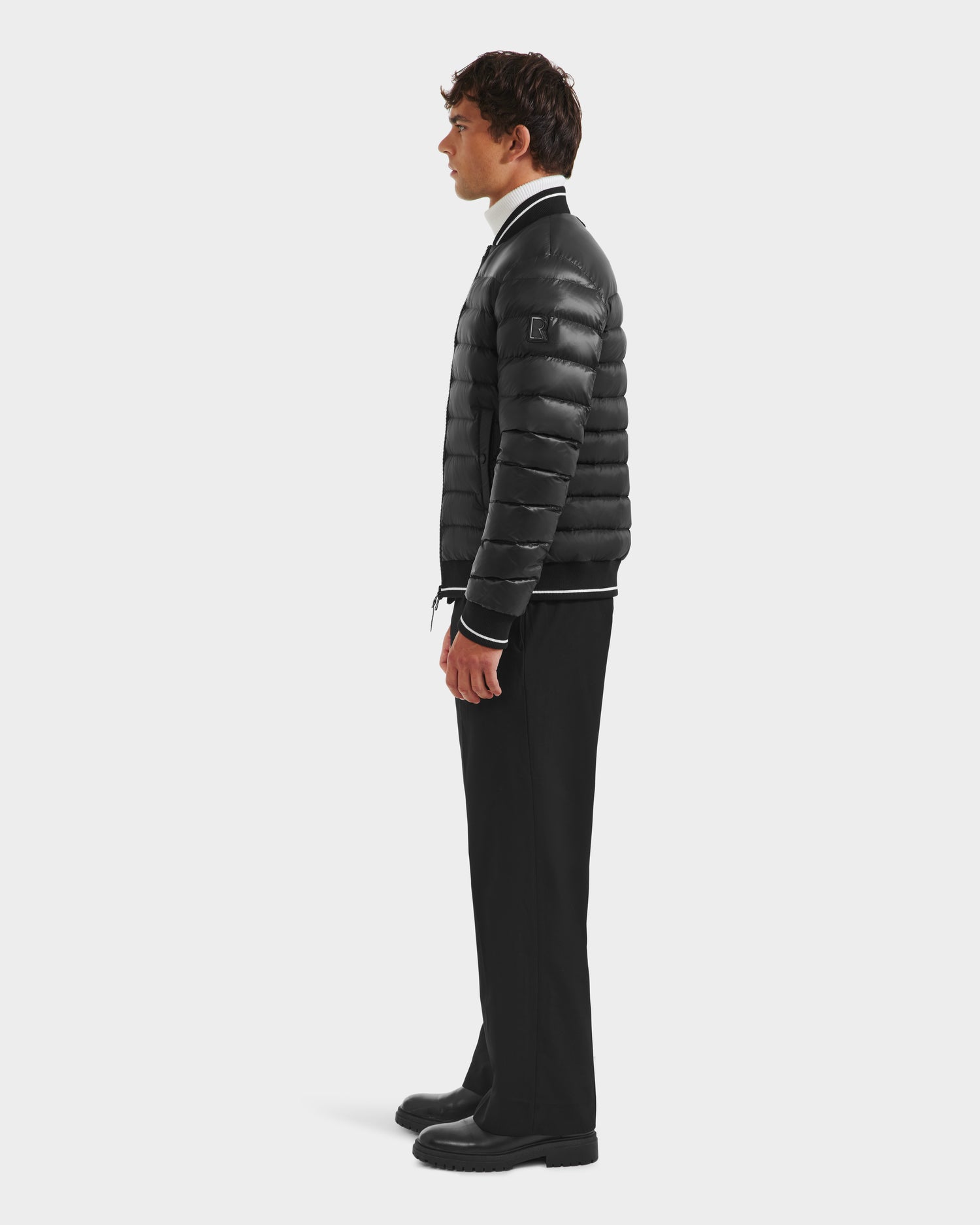A man stands in profile against a white background, wearing the Rudsak JEREMY - 6125025 Black down bomber jacket with black trousers, black shoes, and a white shirt—a perfect minimalist look.
