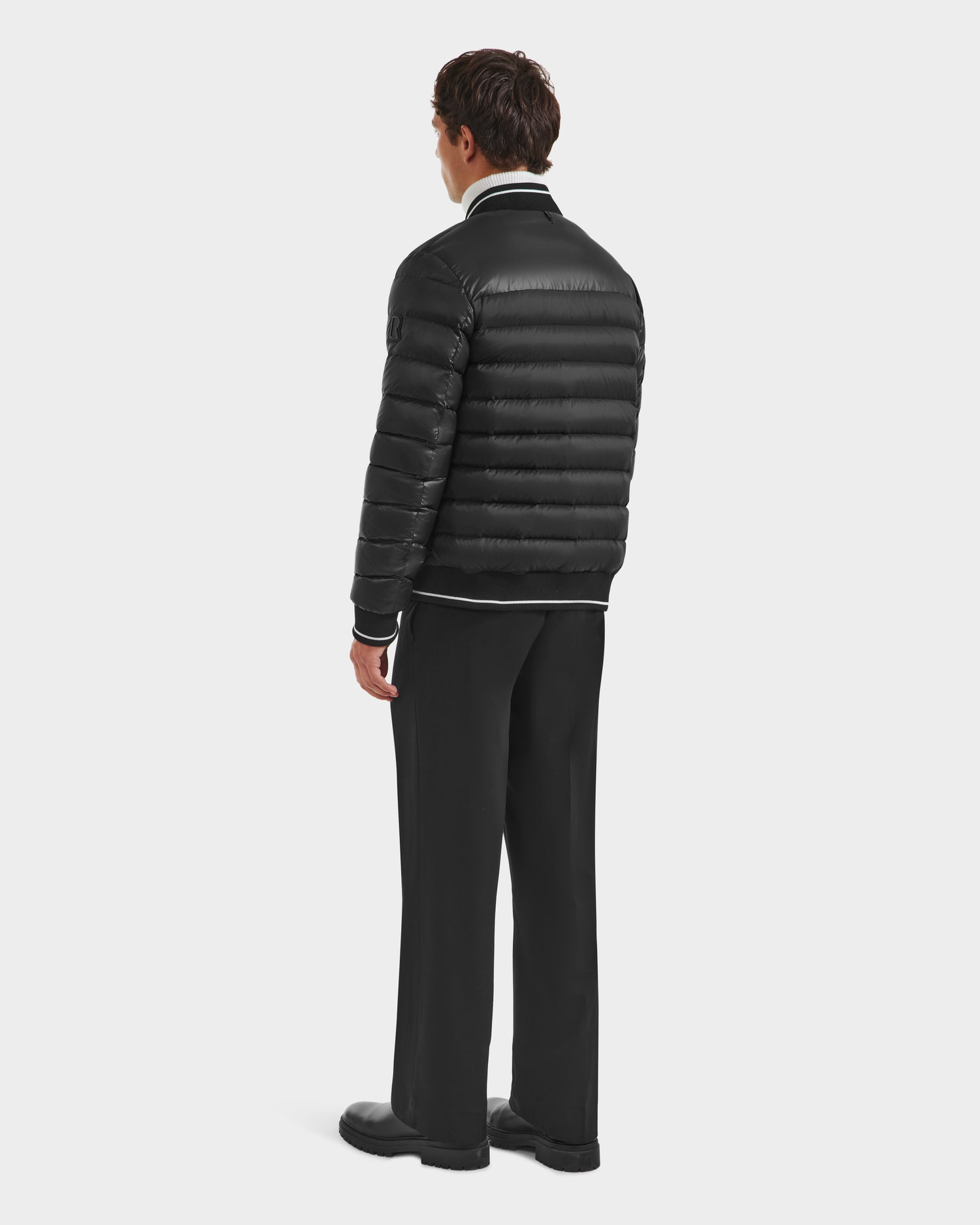 A man faces away wearing the Rudsak JEREMY - 6125025 Black men’s down bomber jacket, paired with black pants and shoes against a white background—an ideal choice for minimalist wardrobes.